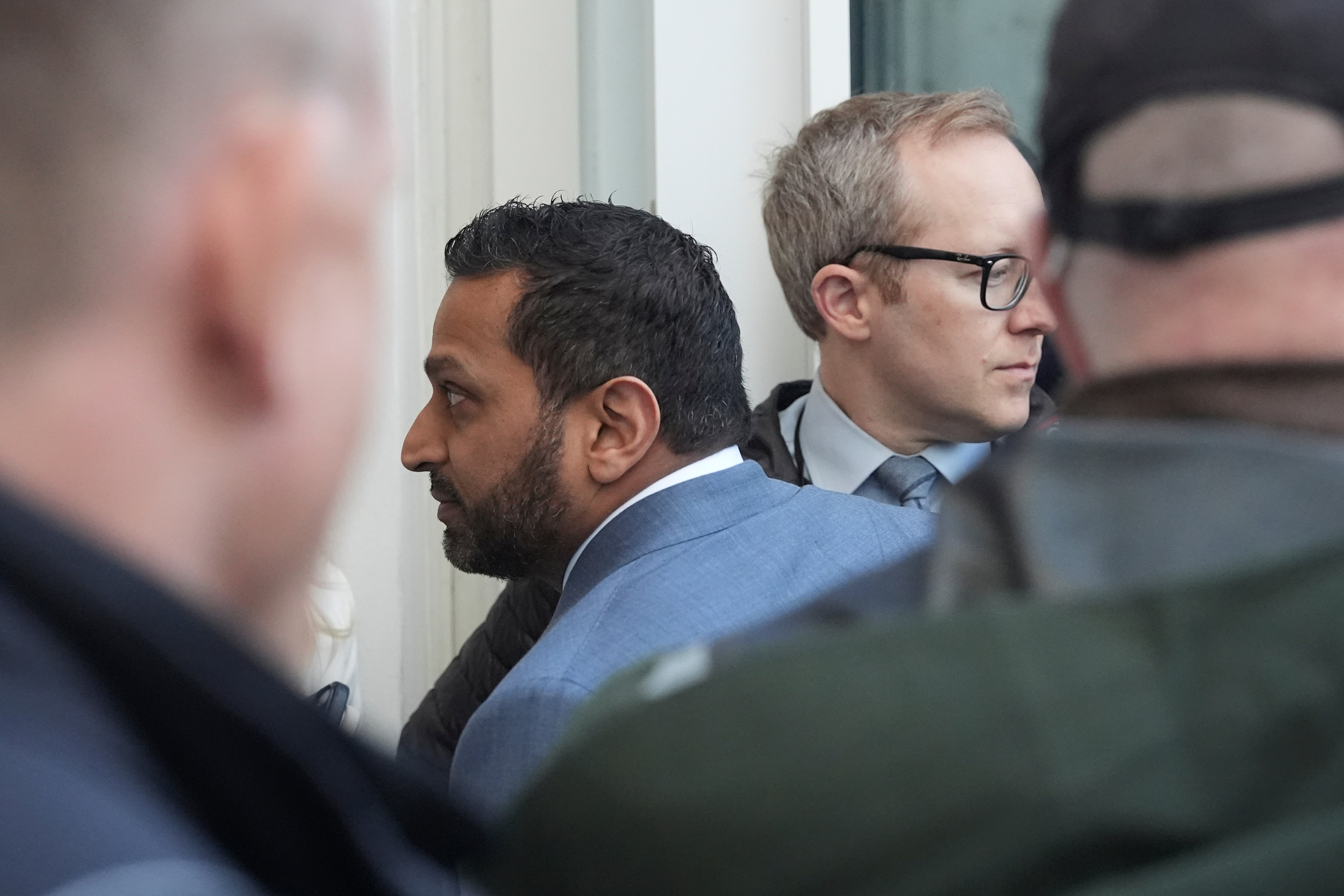 To menn i dress står i en folkemengde og ser i forskjellige retninger. Fokus er på dem, omgitt av delvis synlige mennesker