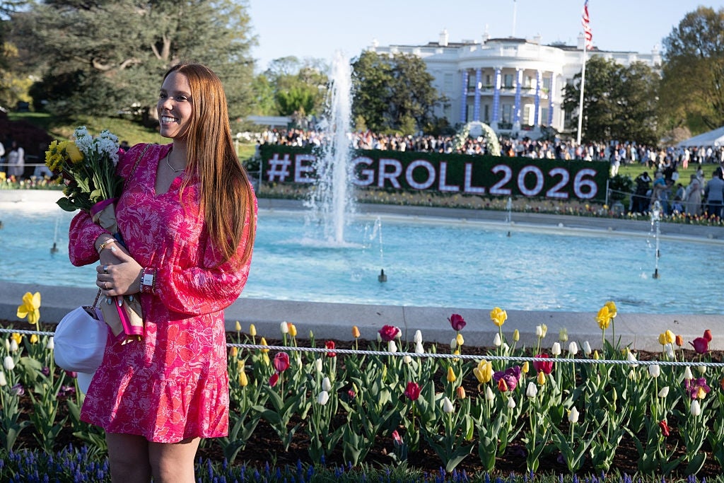 Person i en blomsterkjole holder blomster og smiler foran en fontene. Det hvite hus og "#EggRoll2026" er i bakgrunnen