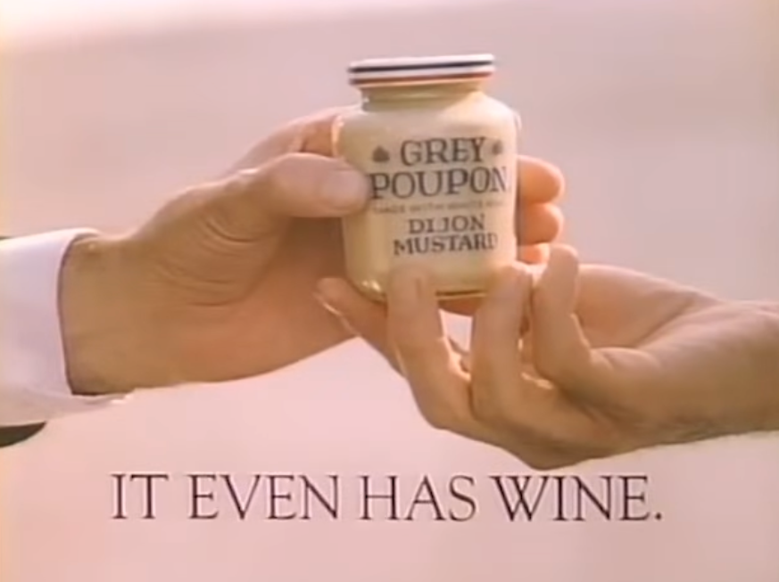 Person hands a jar of Grey Poupon Dijon Mustard to another. Text beneath says, "It Even Has Wine."