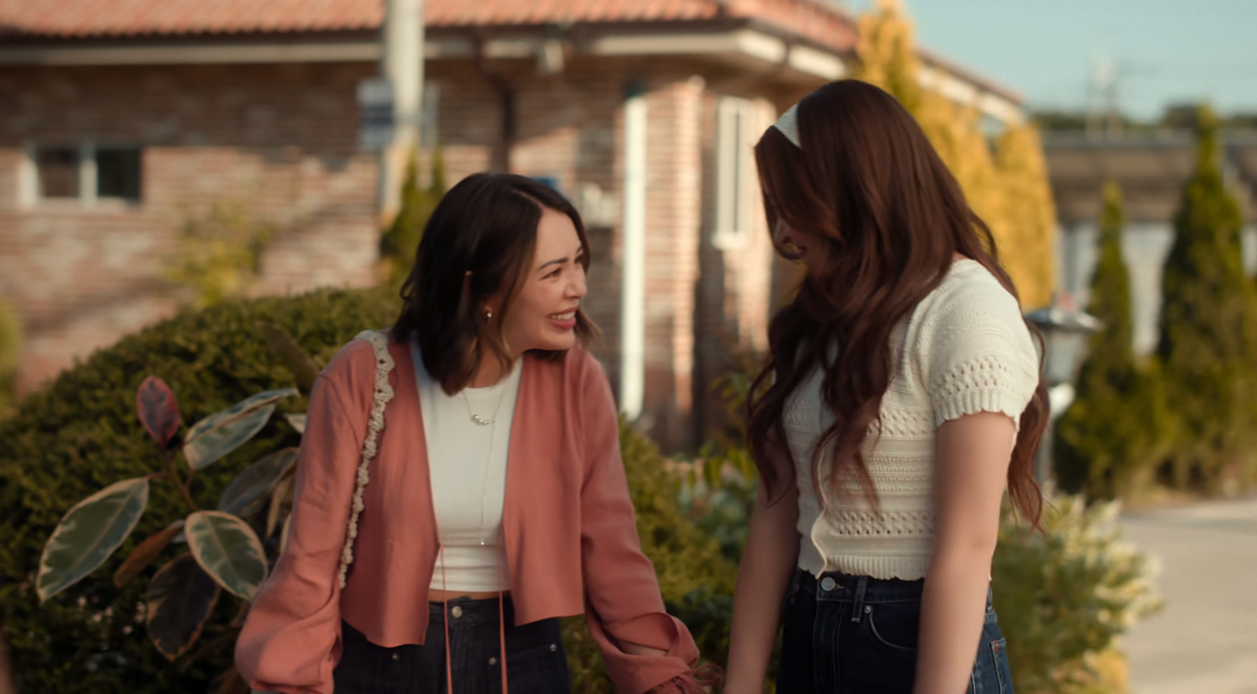 Two women smiling and talking outside, 1 successful a pinkish cardigan and achromatic top, the different successful a achromatic knit top, some wearing acheronian pants