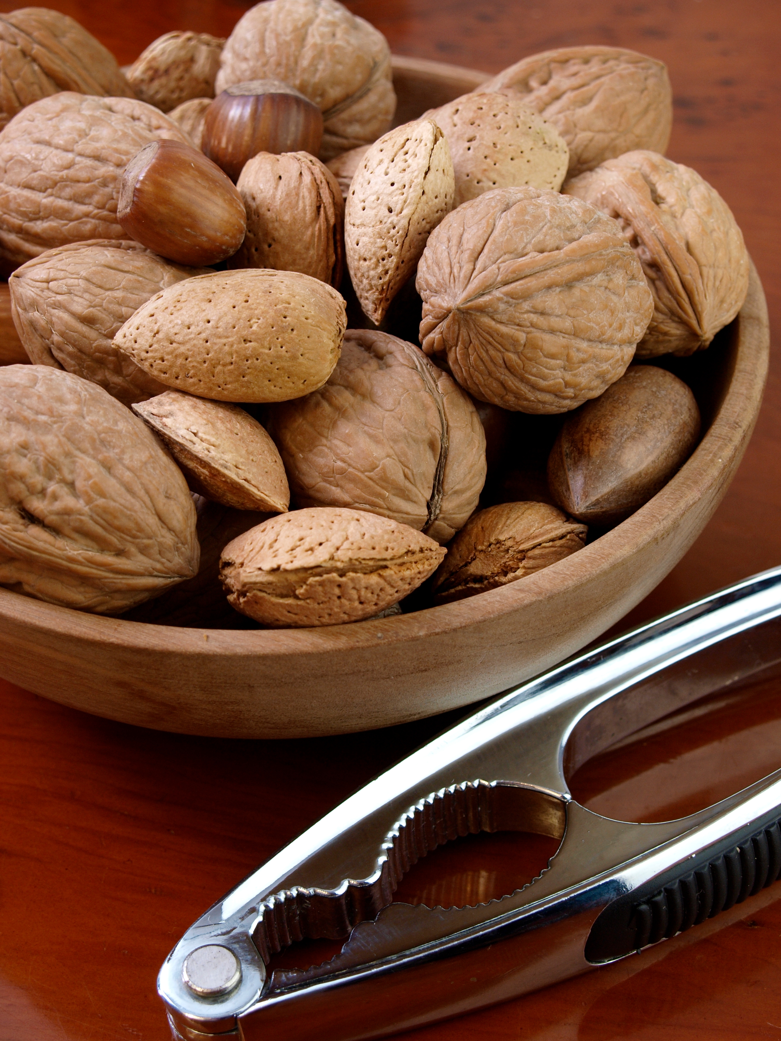 A woody vessel filled with assorted unshelled nuts, including walnuts and almonds, accompanied by a metallic nutcracker connected a table