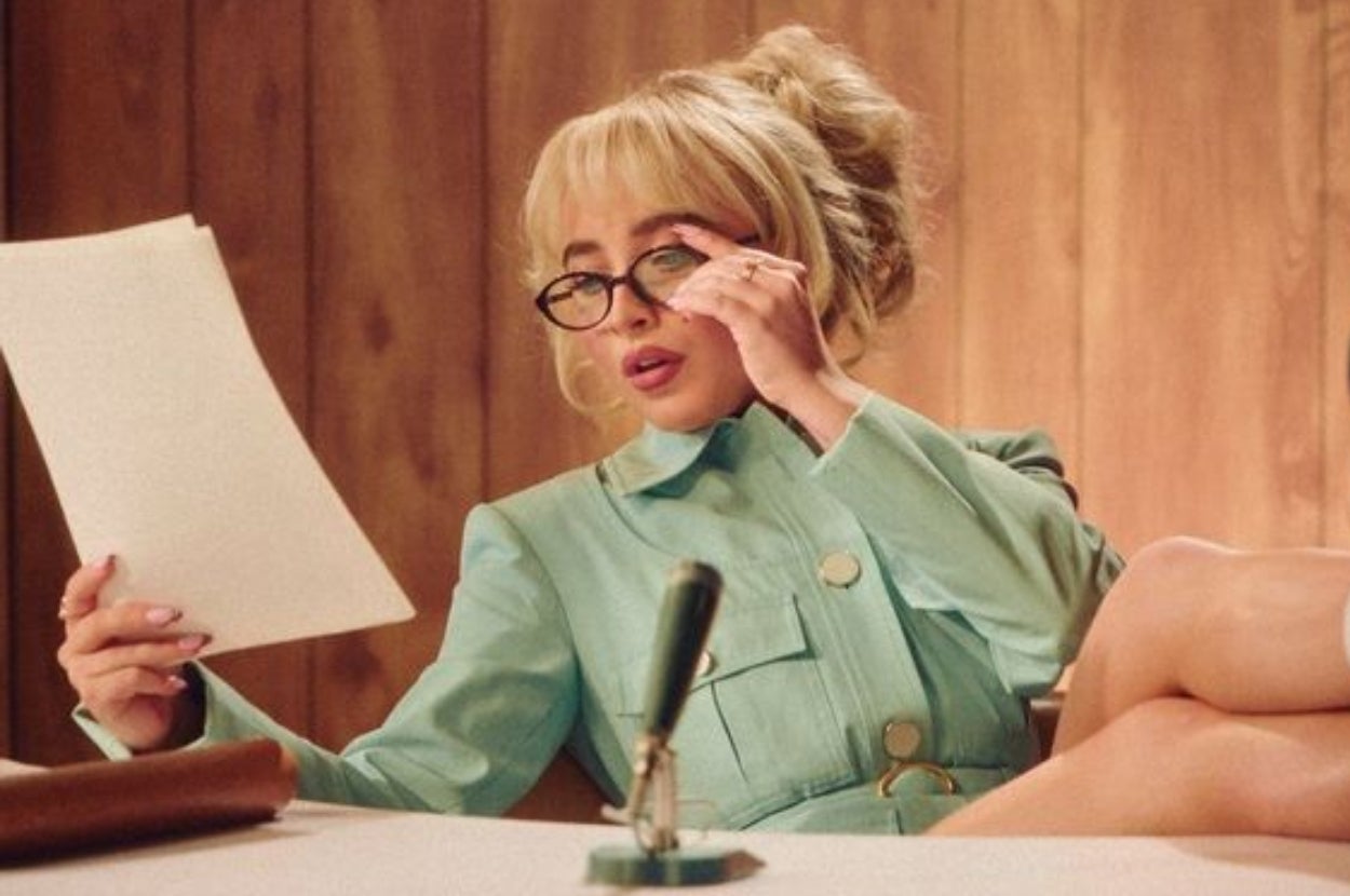Person with blonde hair and glasses reads a document at a desk, dressed in a retro-style outfit