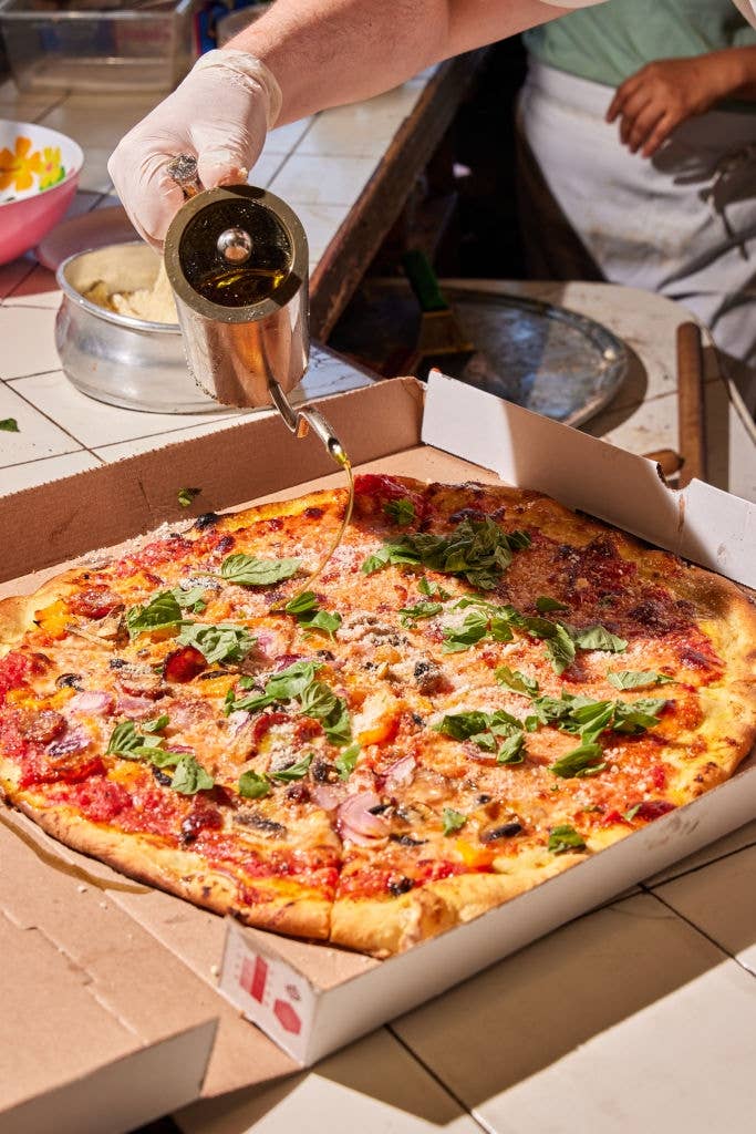 Person pouring oil over a pizza in a takeout box, topped with basil, mushrooms, and other ingredients
