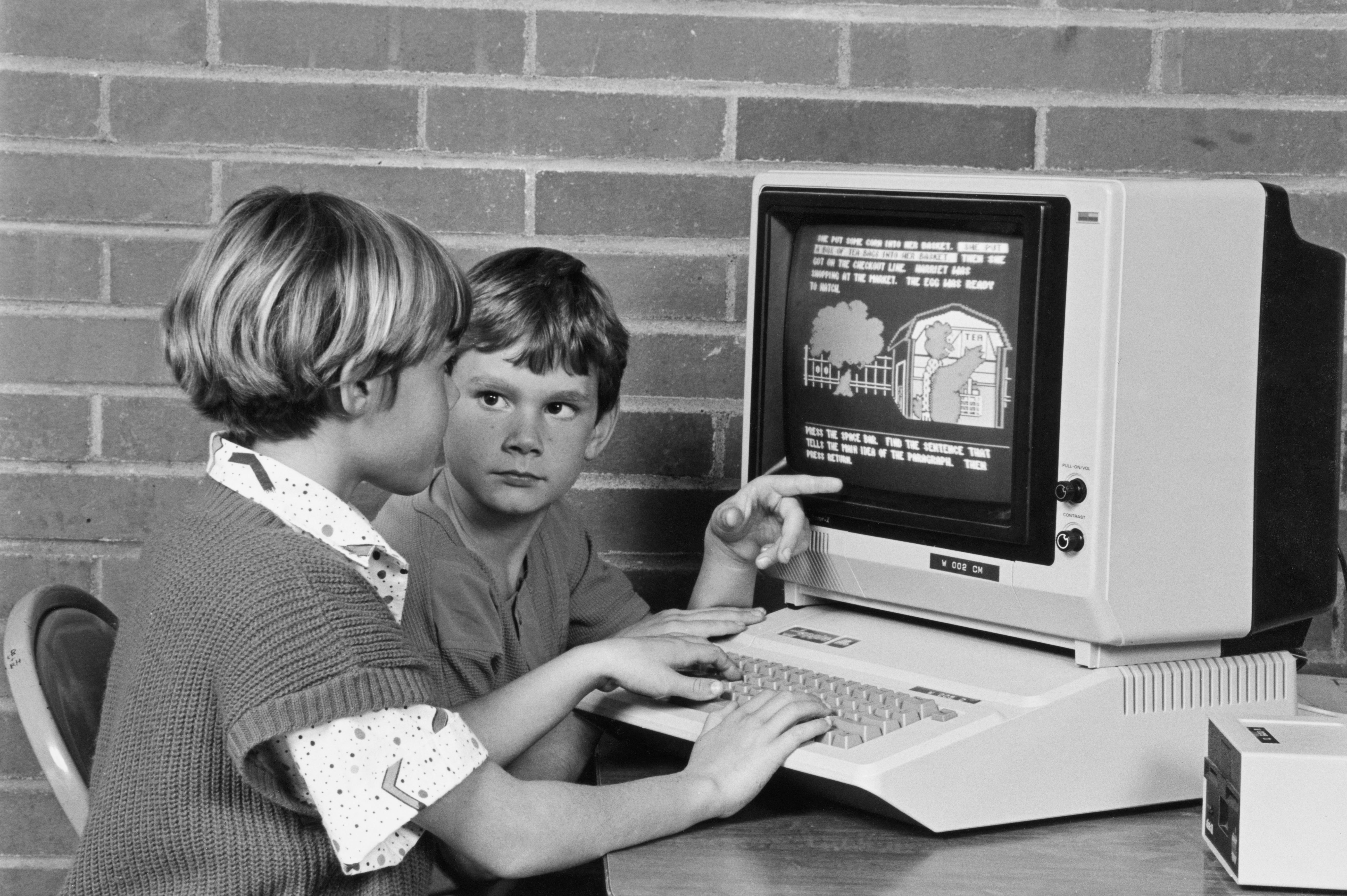 Two children interact with an Apple II computer, 1 typing and the different pointing astatine the substance displayed connected the screen, successful a schoolroom setting