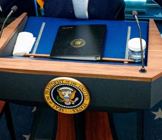 USAs president på et pressekonferansepodium, med tjenestemenn og pressemedlemmer i rommet, diskuterer aktuelle hendelser, en pil peker mot baksiden av håret hans