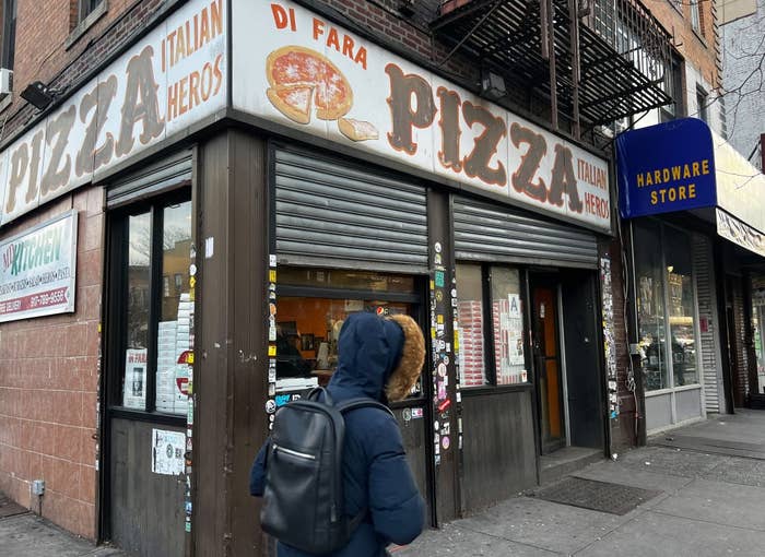 Person walking past Di Fara Pizza storefront with its signage visible; adjacent is a hardware store