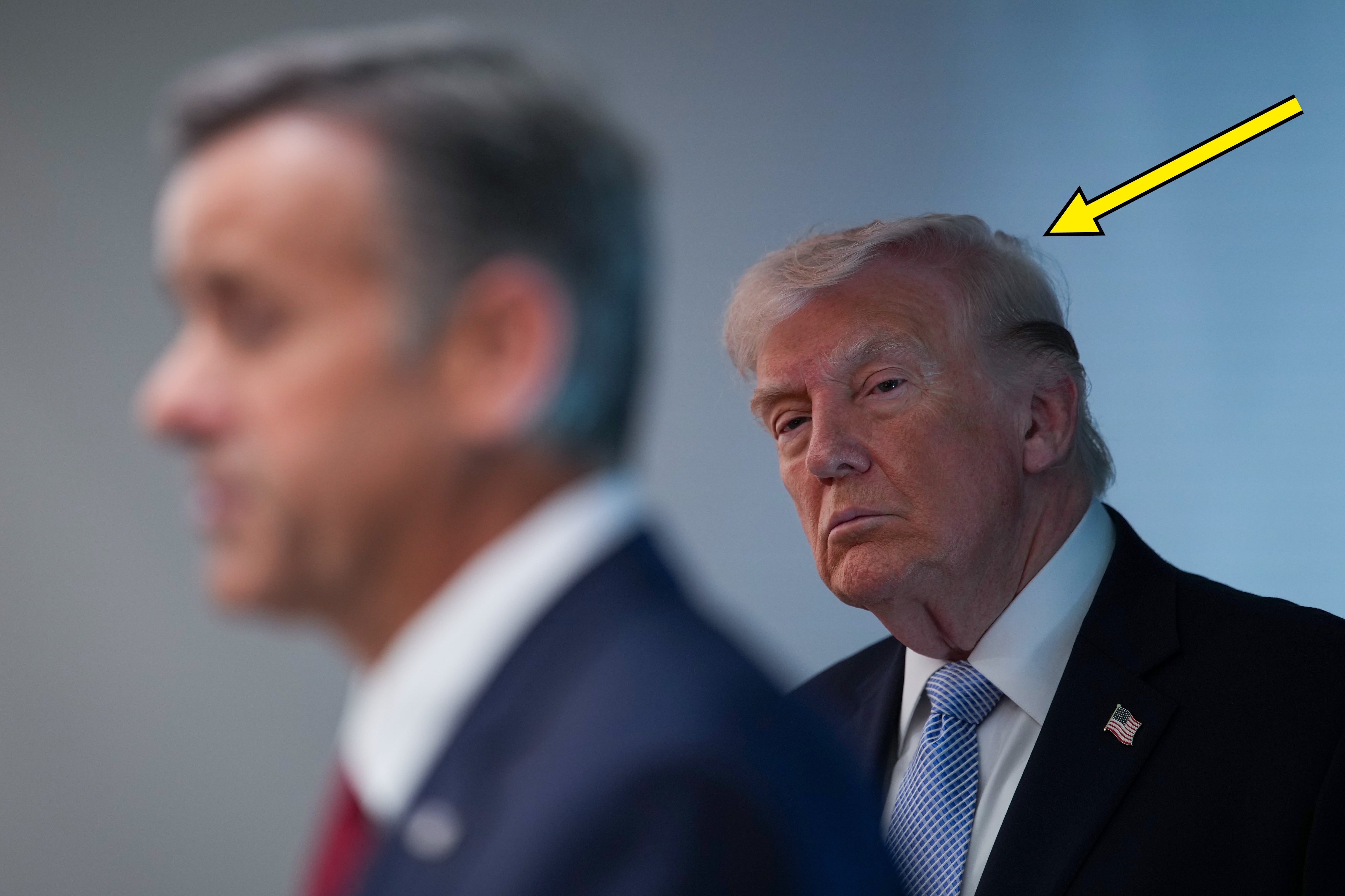 Donald Trump står i bakgrunnen i fokus mens en uskarp mann snakker ved et podium, en pil peker på det rotete håret hans