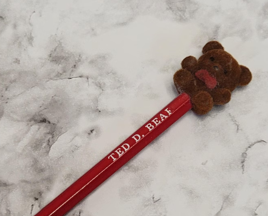 Fuzzy teddy carnivore pencil topper connected a reddish pencil, labeled "Ted D. Bear," against a marble background
