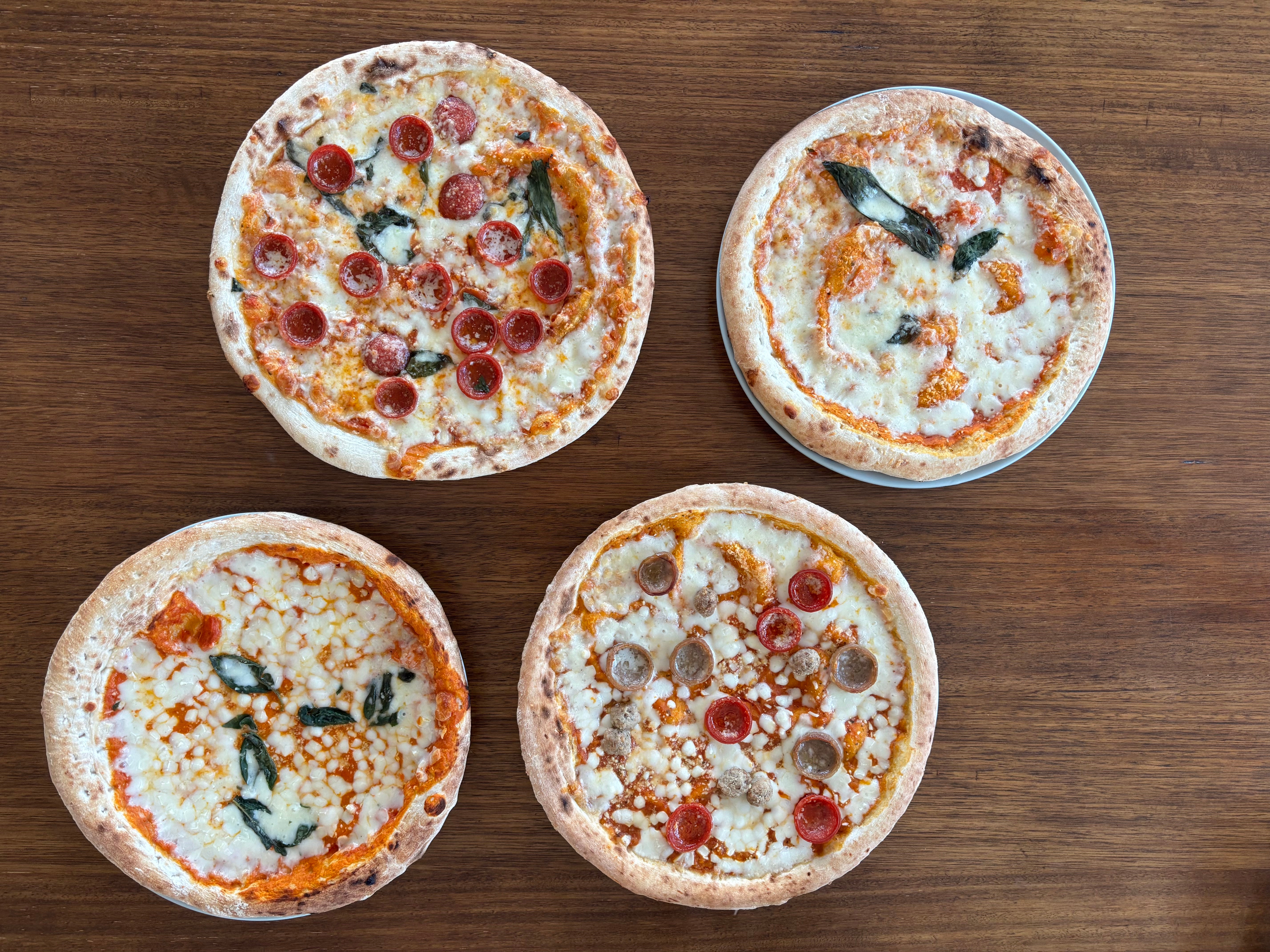 Four pizzas on a wooden table, each topped with various ingredients including pepperoni, sausage, and basil