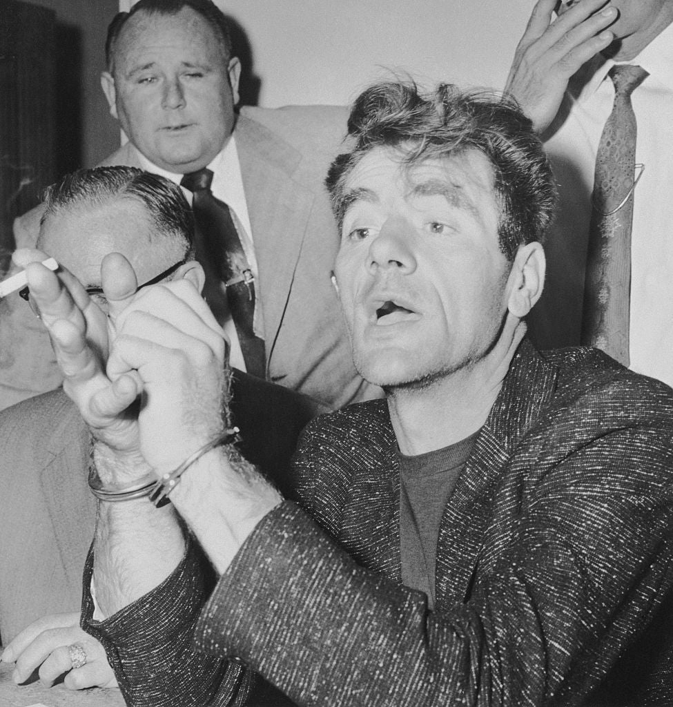 A man in handcuffs speaks animatedly at a press event, with others looking on
