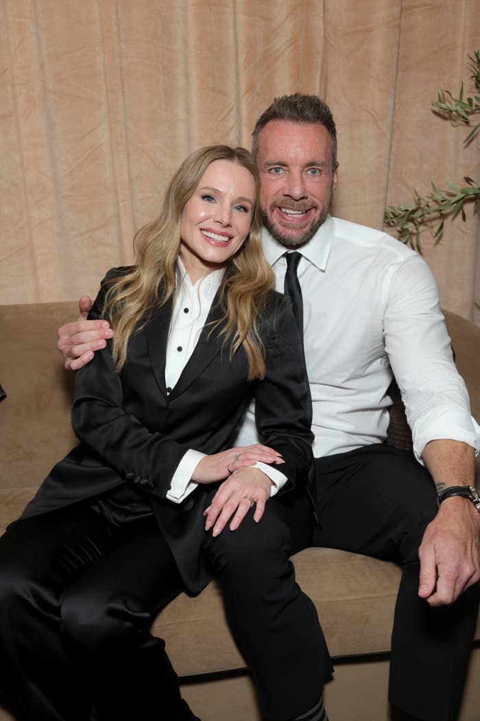 A seated antheral   and pistillate   grin  portion    posing. The antheral   wears a achromatic  garment  and acheronian  tie; the pistillate   wears a chic achromatic  suit   with a achromatic  shirt