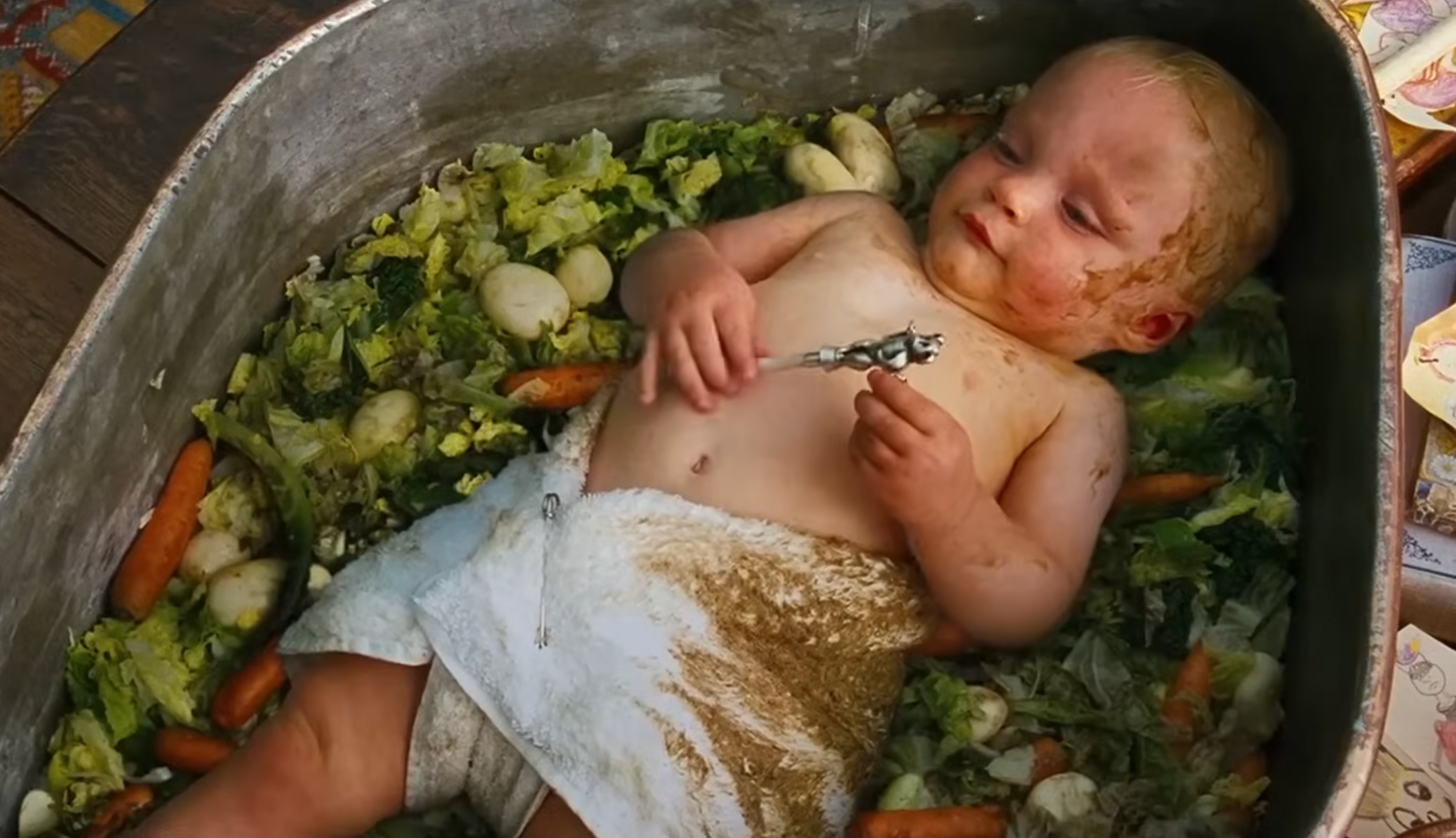 Baby laying successful a tub of vegetables, playing with a utensil, spoofing a cooking cookware scene