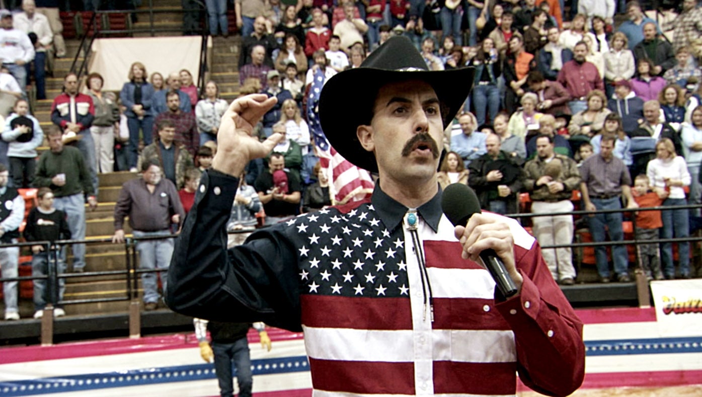 Man successful cowboy chapeau with mustache holds microphone, wearing American emblem shirt, speaking successful crowded arena, U.S. emblem disposable behind