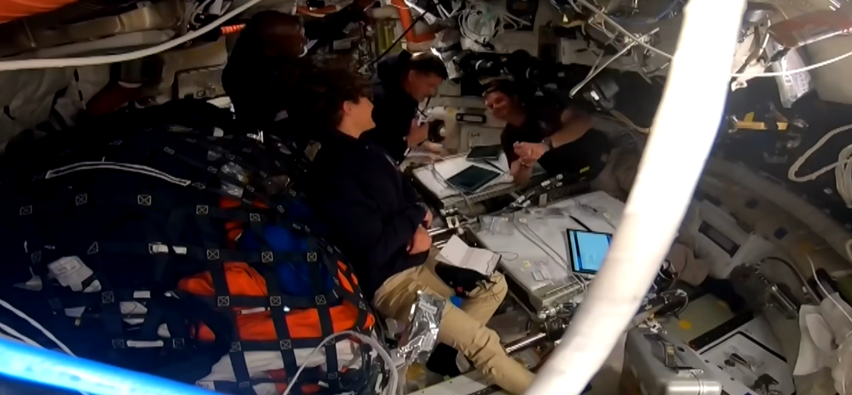 Astronauter i en romfartøyshytte, engasjert i aktiviteter med nettbrett og bærbare datamaskiner, omgitt av utstyr og sikrede vesker