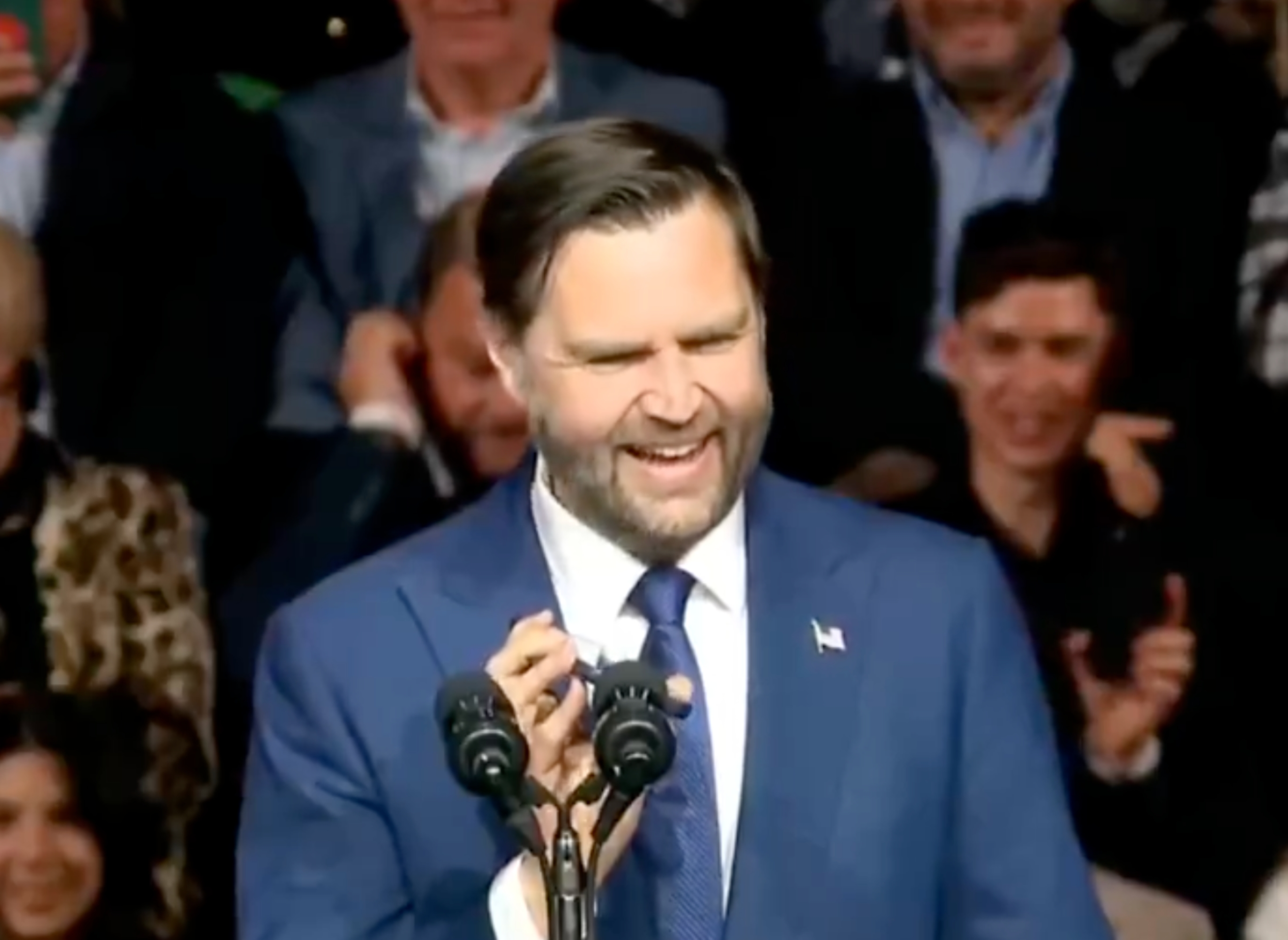 A idiosyncratic successful a suit is speaking astatine a podium with a microphone, smiling. An assemblage is disposable successful the background, immoderate taking photos