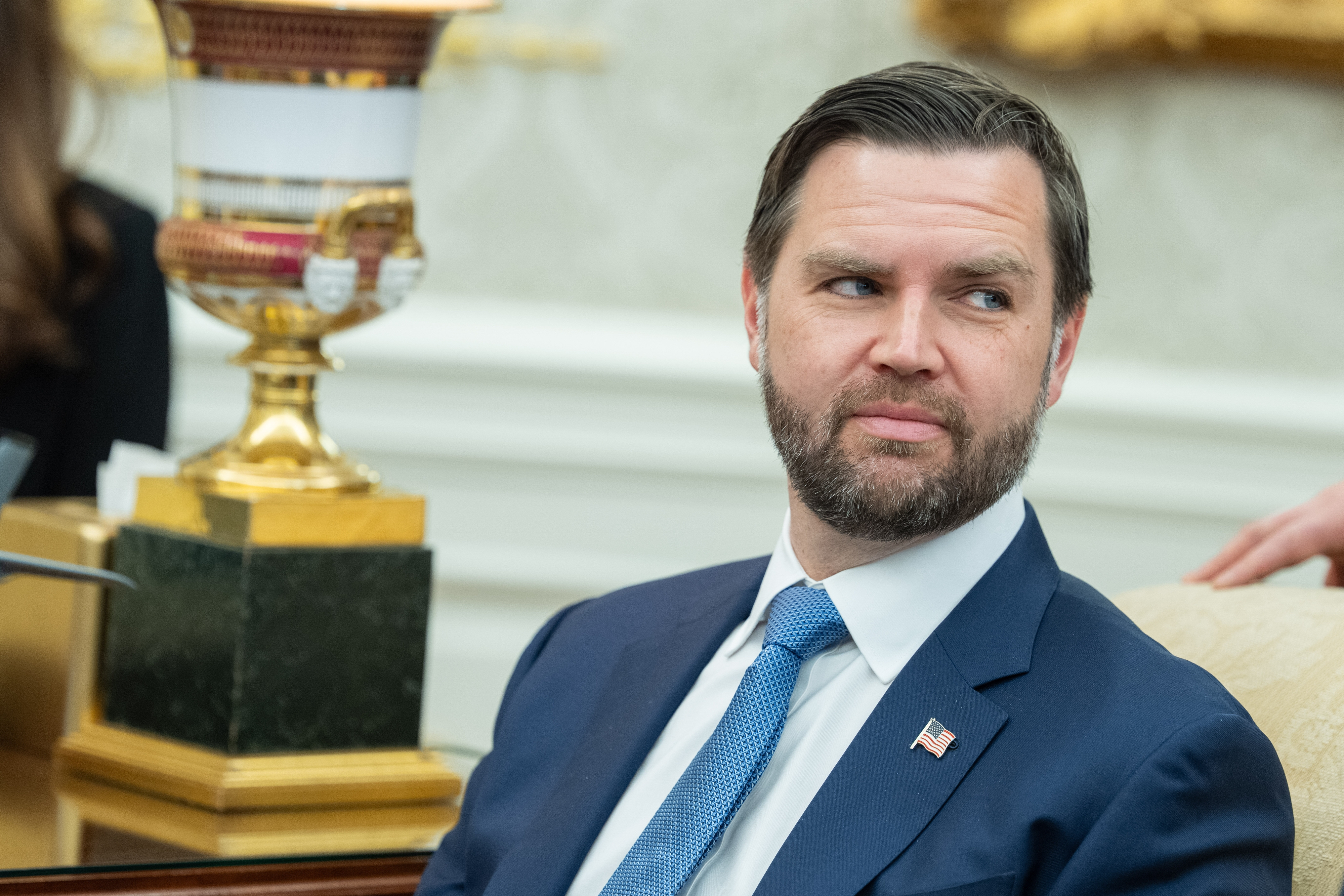 A antheral successful a suit and necktie with a tiny U.S. emblem pin sits astatine a ceremonial meeting, looking sideways. A decorative lamp is successful the background