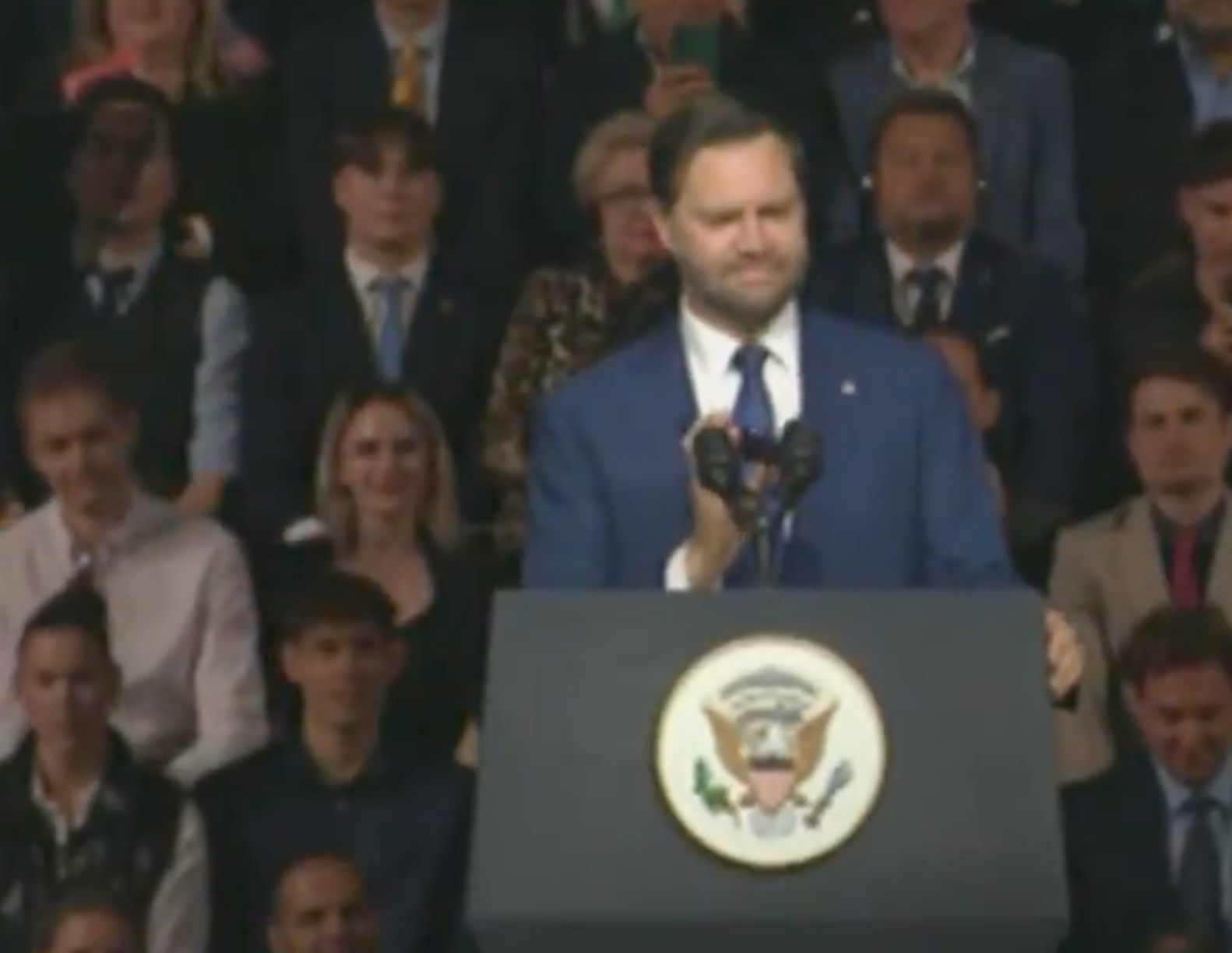 Man successful suit speaking astatine a podium with statesmanlike seal, assemblage blurred successful background