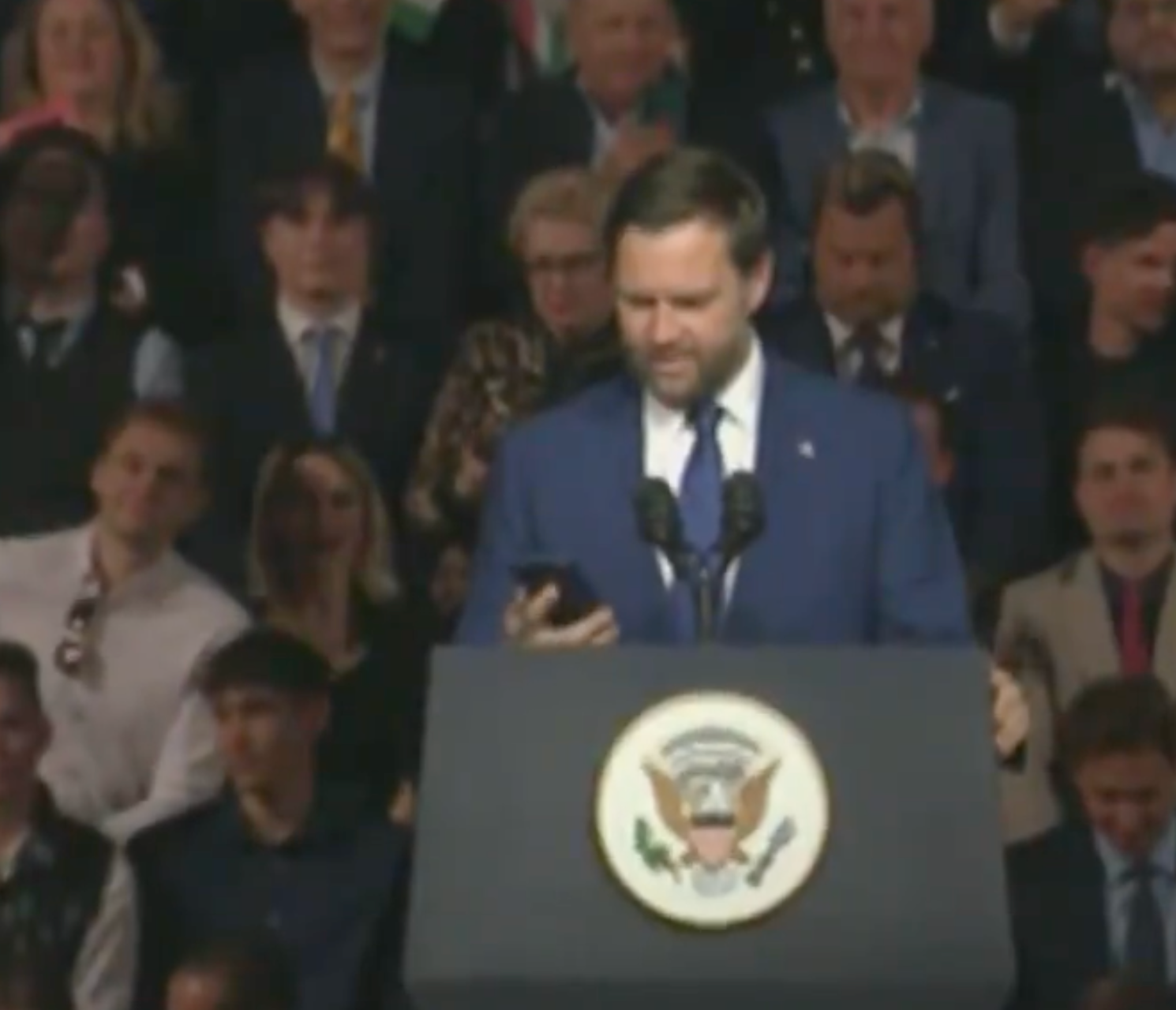 A antheral successful a suit reads from a telephone astatine a podium with the U.S. statesmanlike seal, surrounded by a crowd