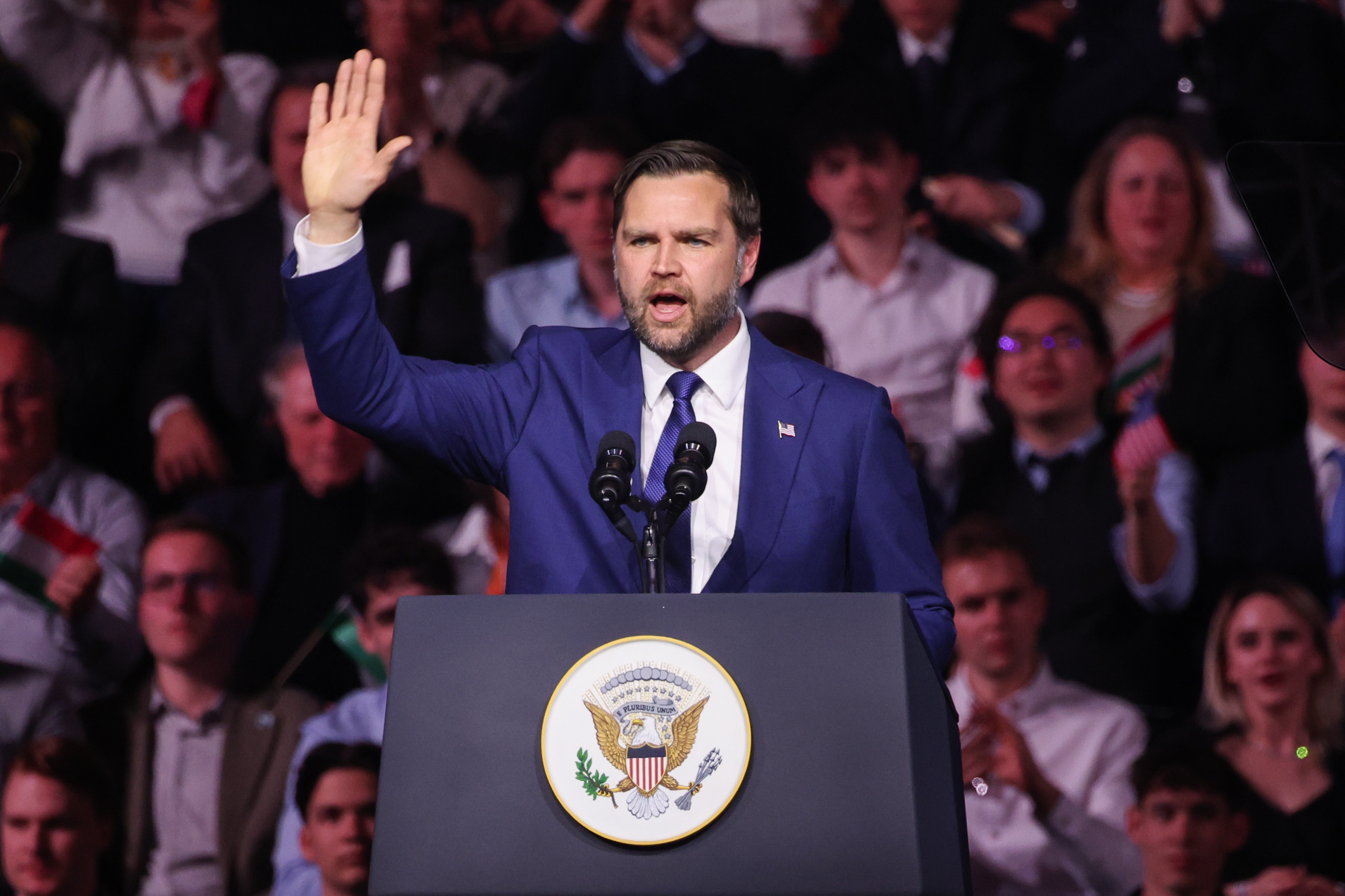 A antheral successful a suit gestures portion speaking astatine a podium with a statesmanlike seal, surrounded by a crowd
