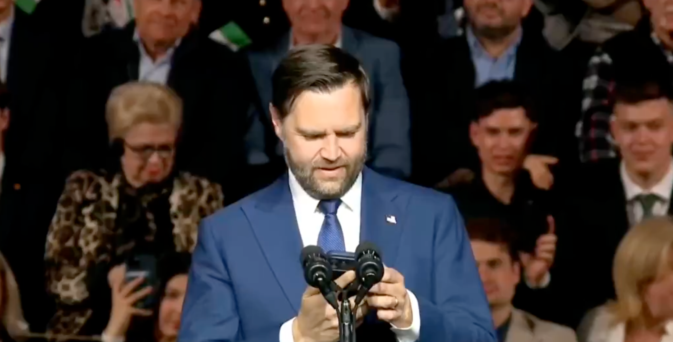 Man successful a suit speaking astatine a podium with a assemblage down him