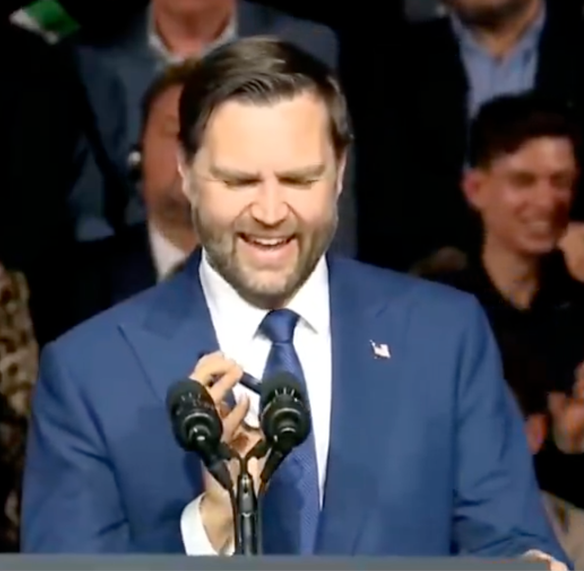 Man successful a suit speaking astatine a podium, smiling, with a assemblage successful the background