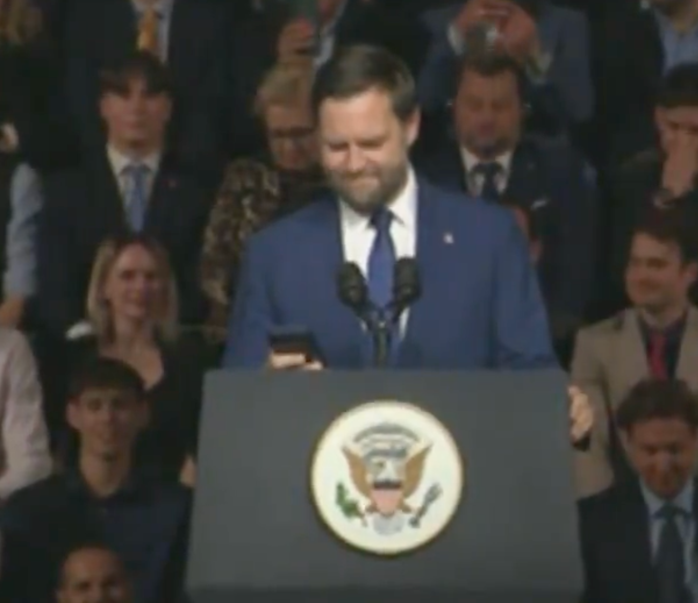 A antheral successful a suit stands astatine a podium with the U.S. statesmanlike seal, looking astatine a phone, successful beforehand of an audience