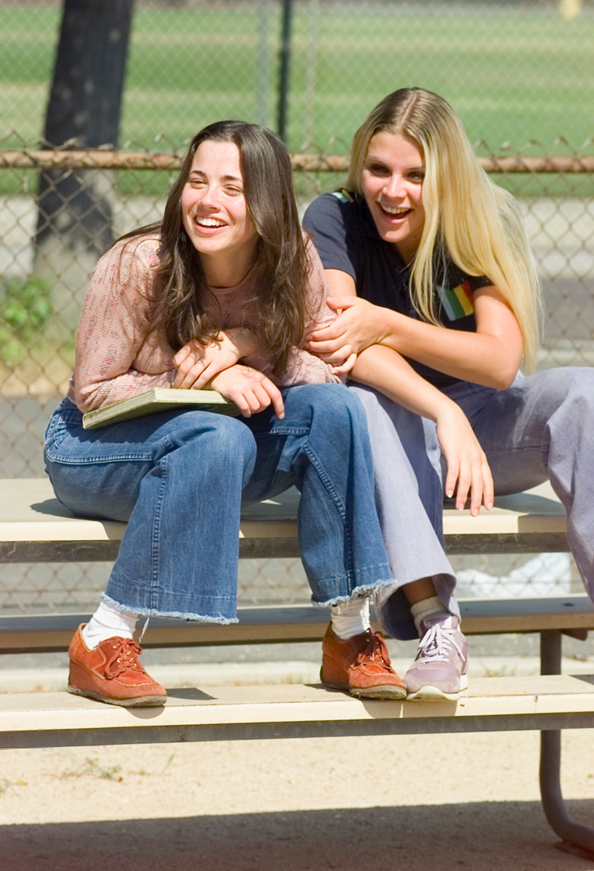 Two people sitting on a bench, smiling joyfully. One wears a patterned top and jeans, the other a striped shirt and flared pants
