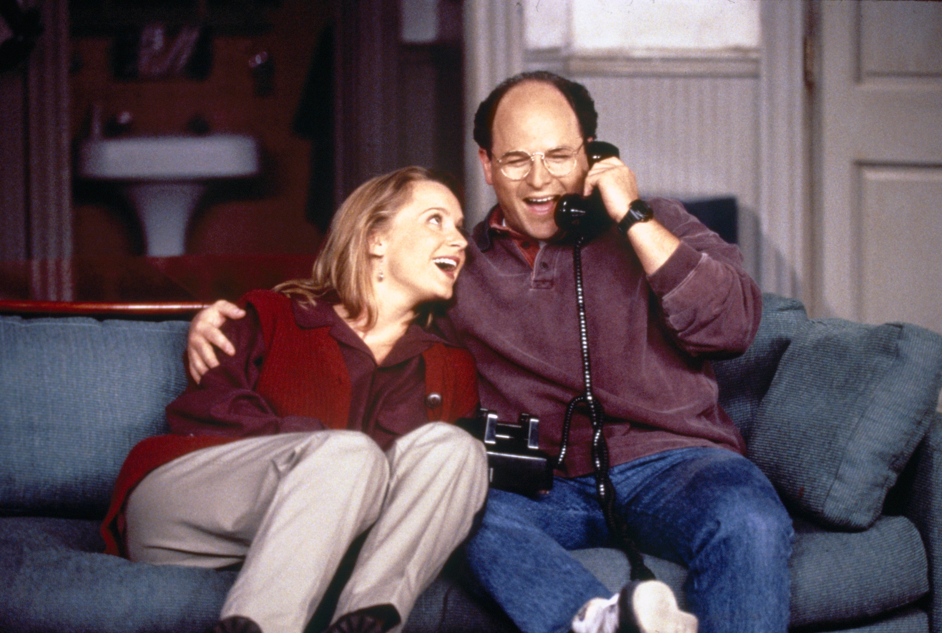 Two people sitting on a couch, laughing while sharing a phone call. One holds a handset and a remote. Casual 90s attire