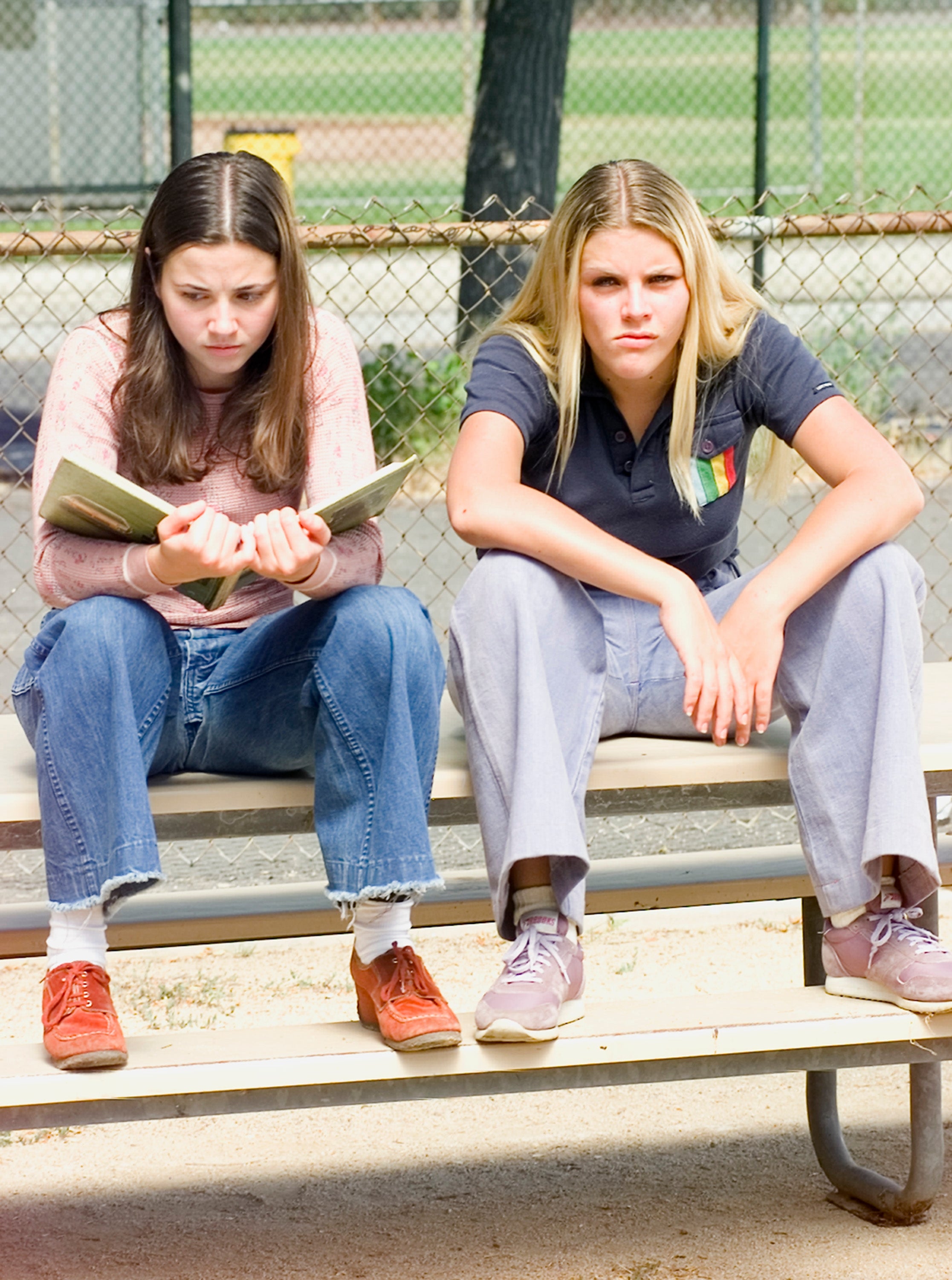 Two people sitting on a bench, one reading a book, both with serious expressions. They are casually dressed in jeans and shirts