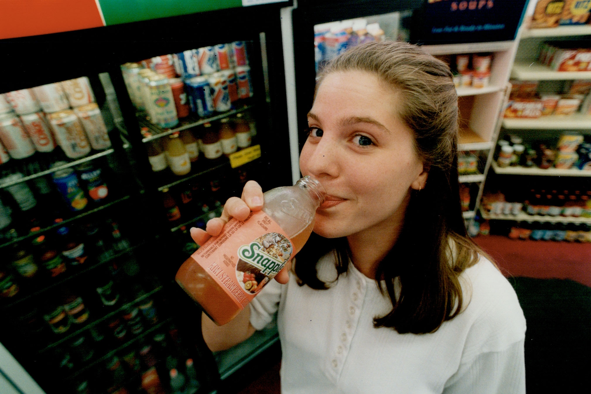Person drinking a Snapple beverage wrong a convenience store. They're facing the camera with a flimsy smile. Shelves of drinks are disposable behind