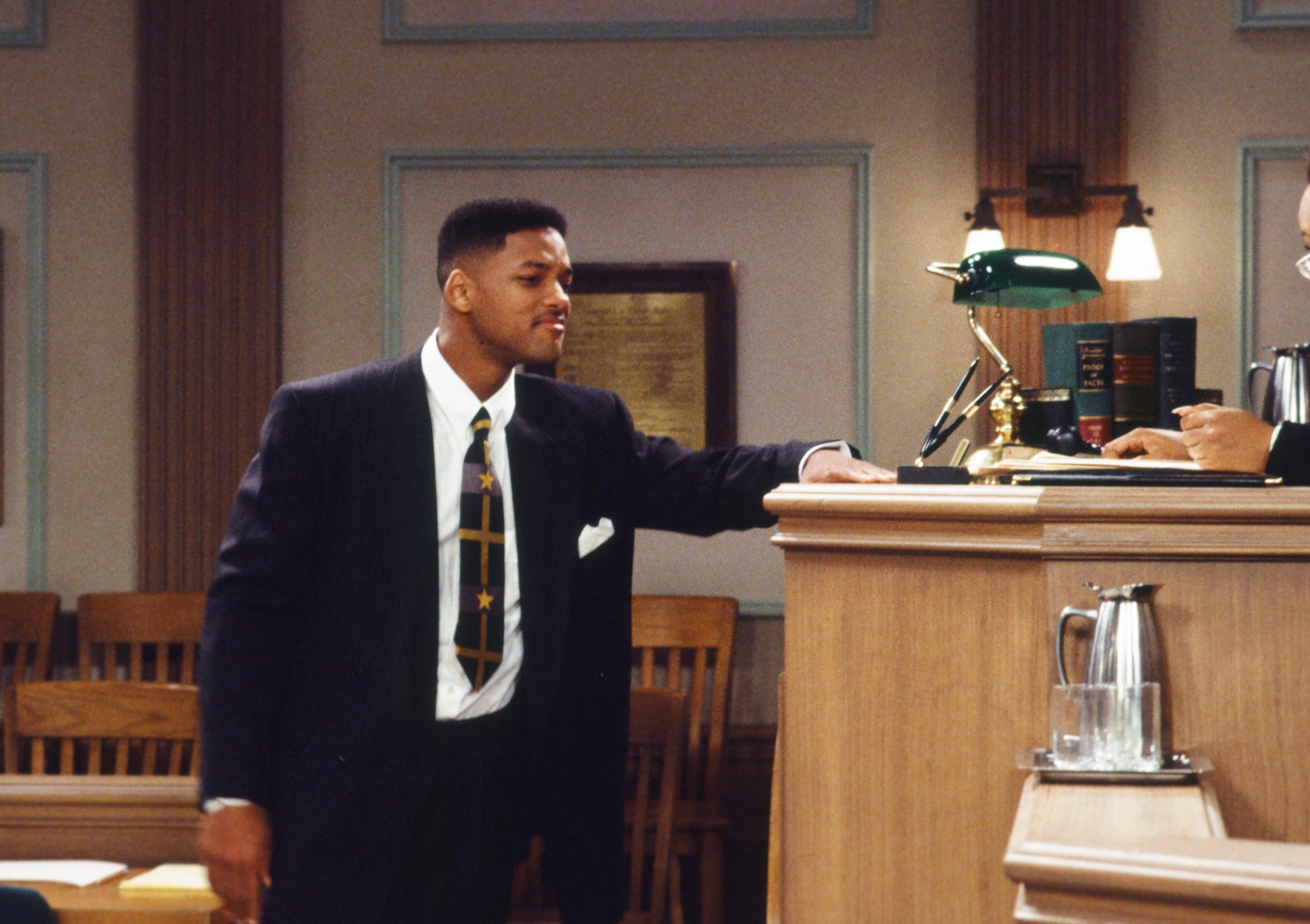 A person in a courtroom setting wearing a stylish suit and patterned tie, speaking with a judge