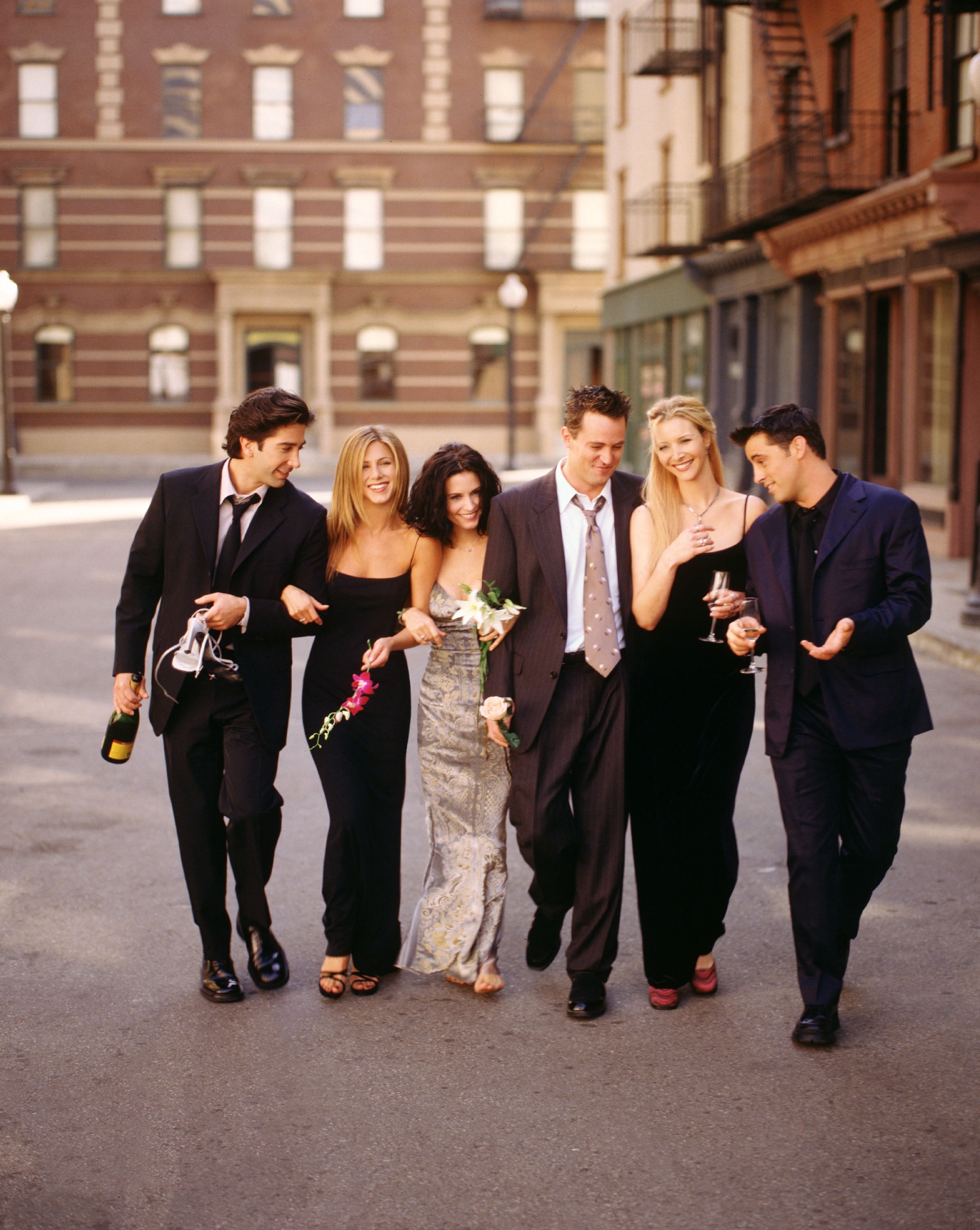 Six people in formal attire strolling down a street, smiling and holding flowers. Their clothing includes suits and elegant dresses