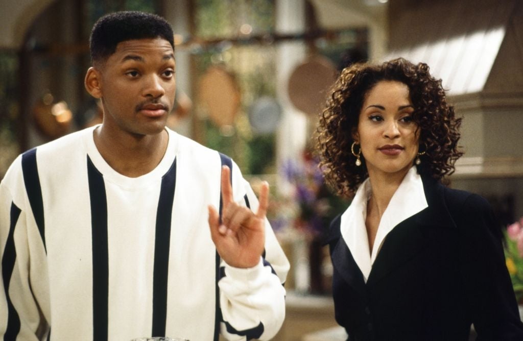 Two actors from a 1990s sitcom scene; one wears a casual shirt, the other a professional outfit. They appear engaged in conversation