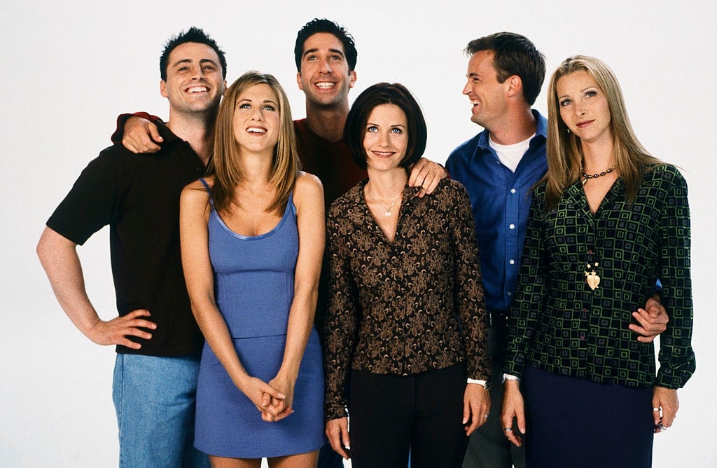 Group of six actors from a popular TV show, standing together and smiling, dressed in casual 90s fashion