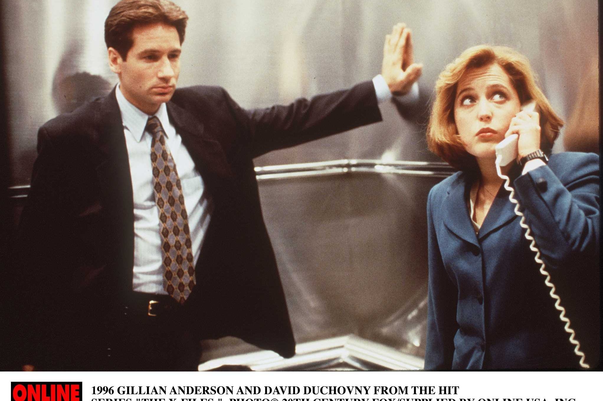 Two people in business attire; one holds a phone, looking up thoughtfully. Scene relates to "The X-Files."