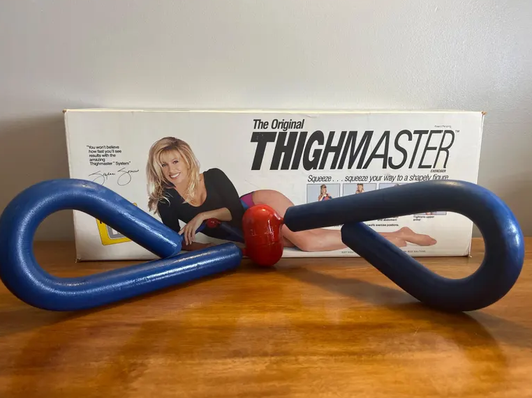 Box with a representation of a personage utilizing a ThighMaster workout tool; the instrumentality is bluish with a reddish center