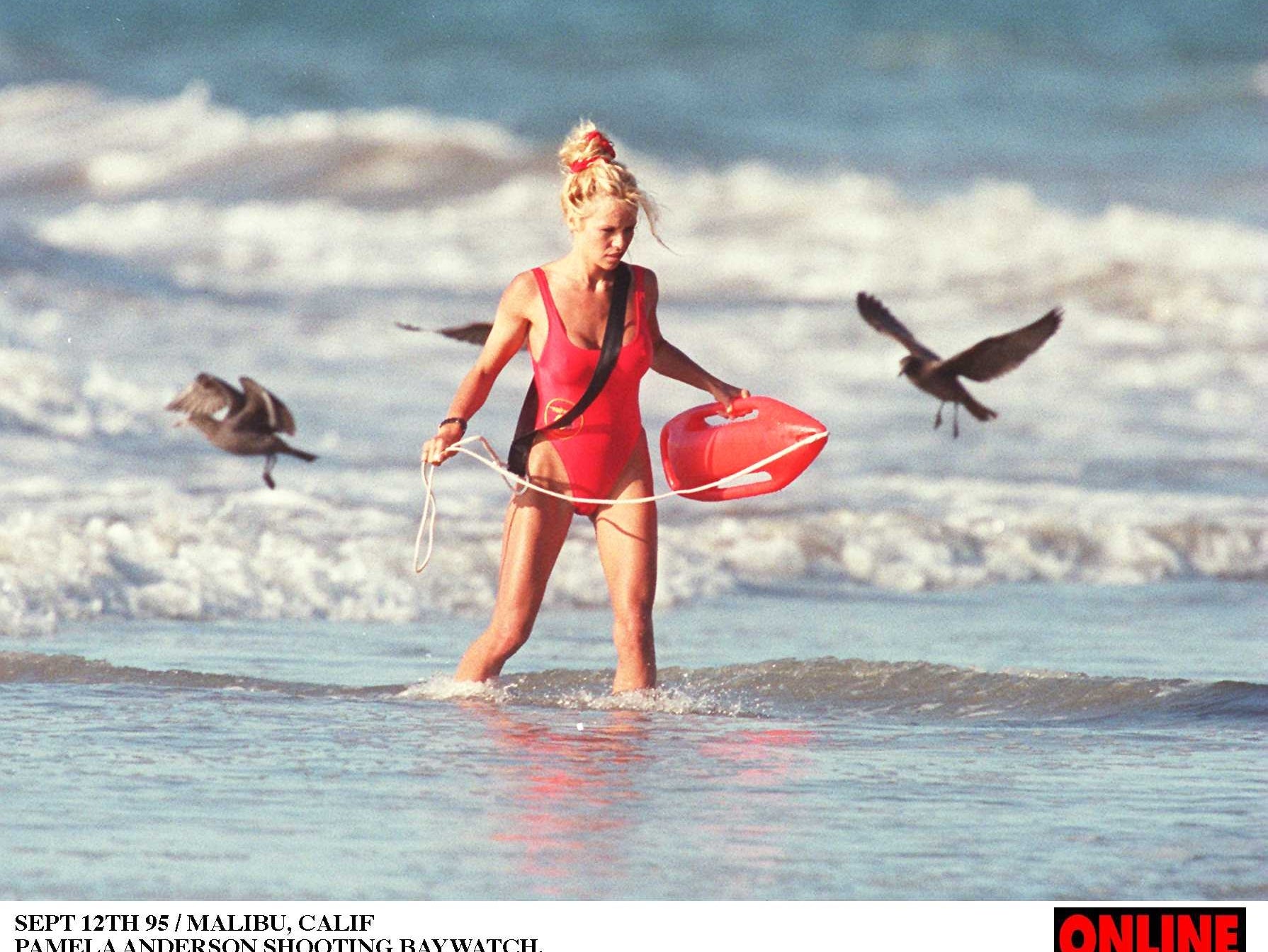 Person in a red one-piece swimsuit walks through water holding a float, surrounded by seagulls, filmed for "Baywatch."