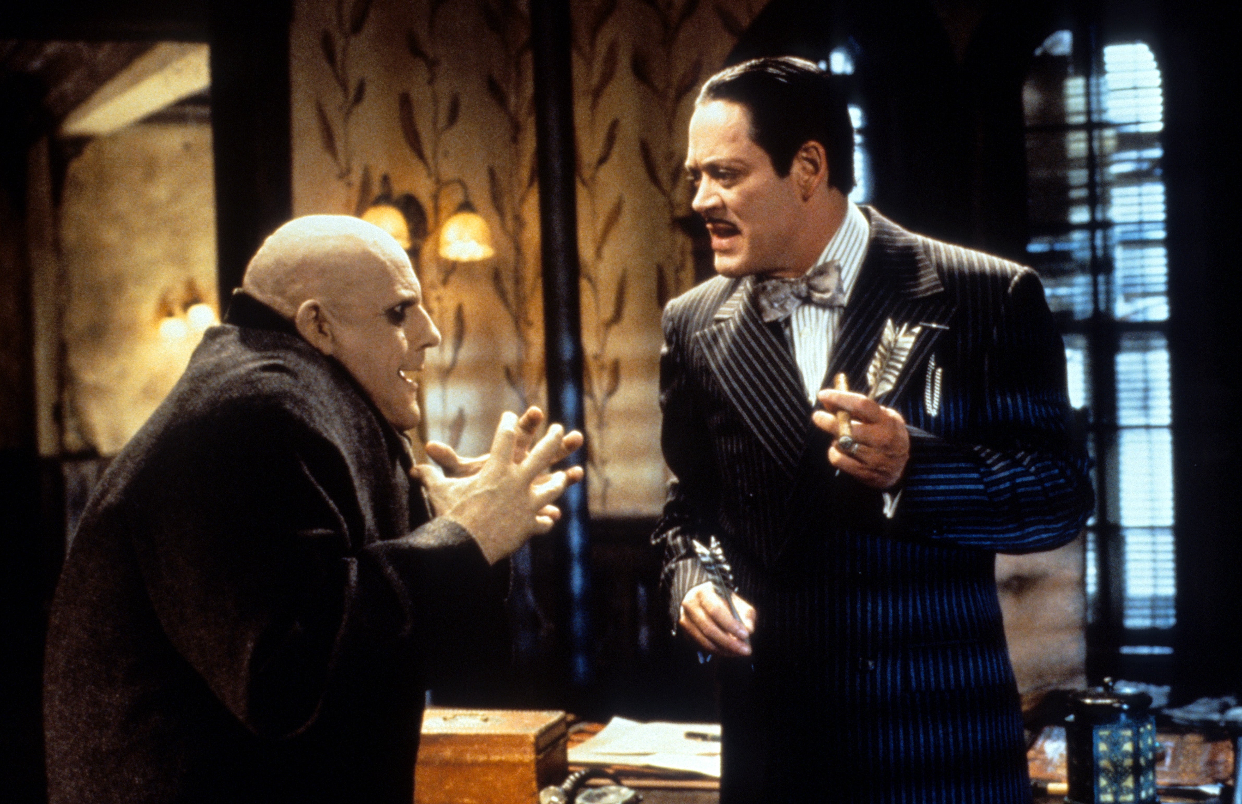  one with a bald head and dark cloak, the other in a pinstripe suit with a bow tie, engaged in conversation
