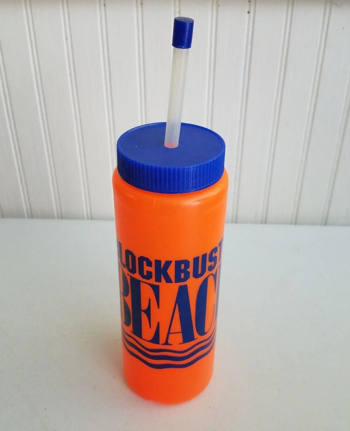 Orange "BLOCKBUSTER BEACH" tumbler with a bluish headdress and straw, placed connected a achromatic surface