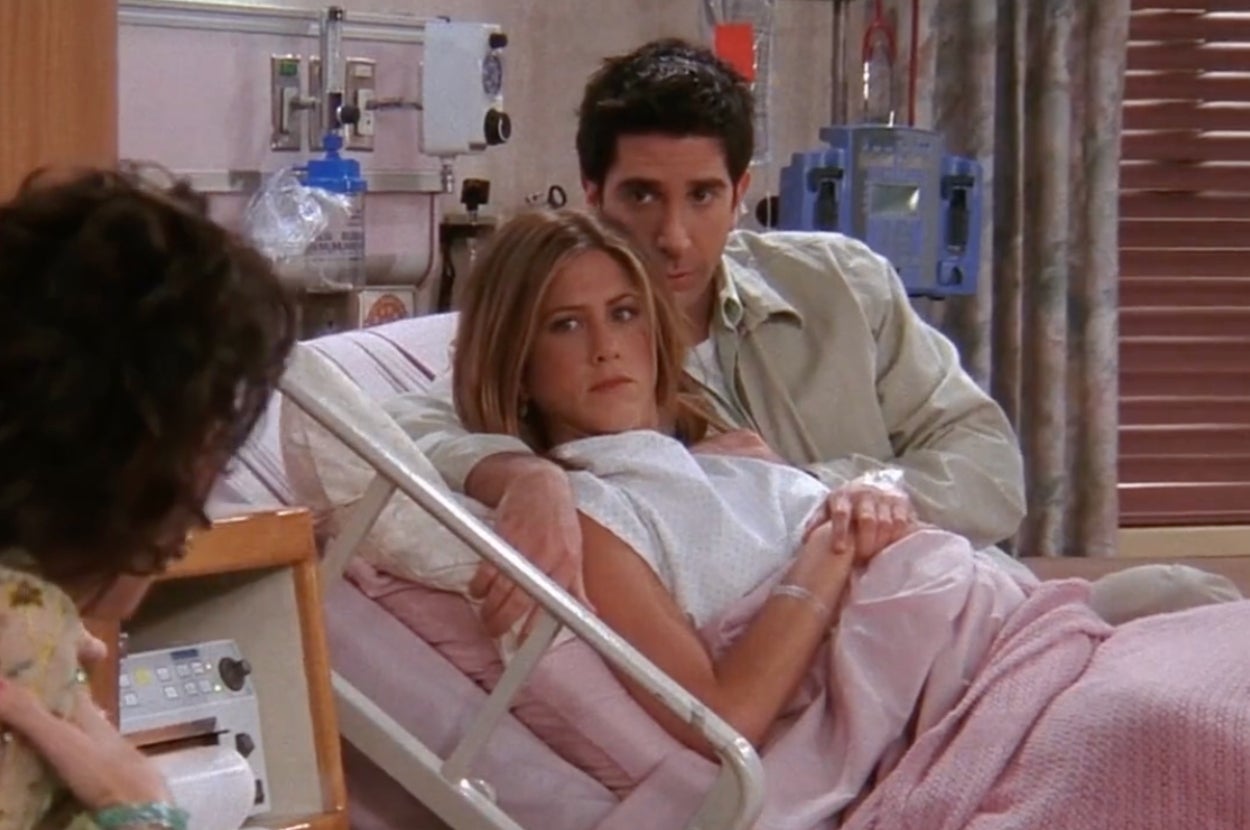 A woman and man are sitting on a hospital bed, with the man supporting the woman. A third person is seated nearby, partially visible