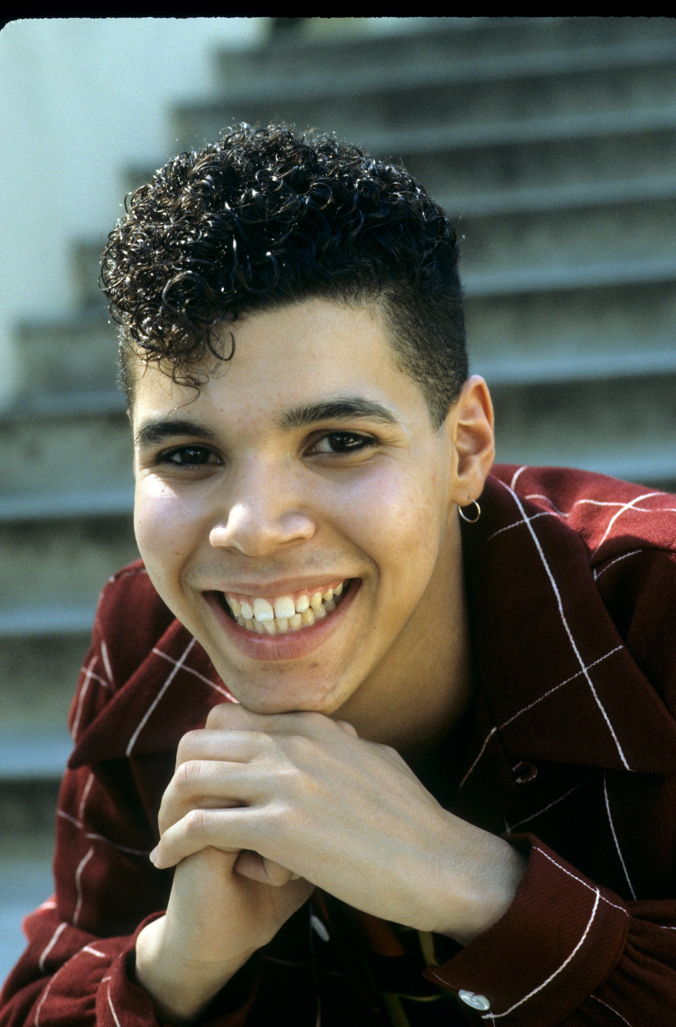 A person with a big smile poses on steps, wearing a plaid shirt with hands under their chin