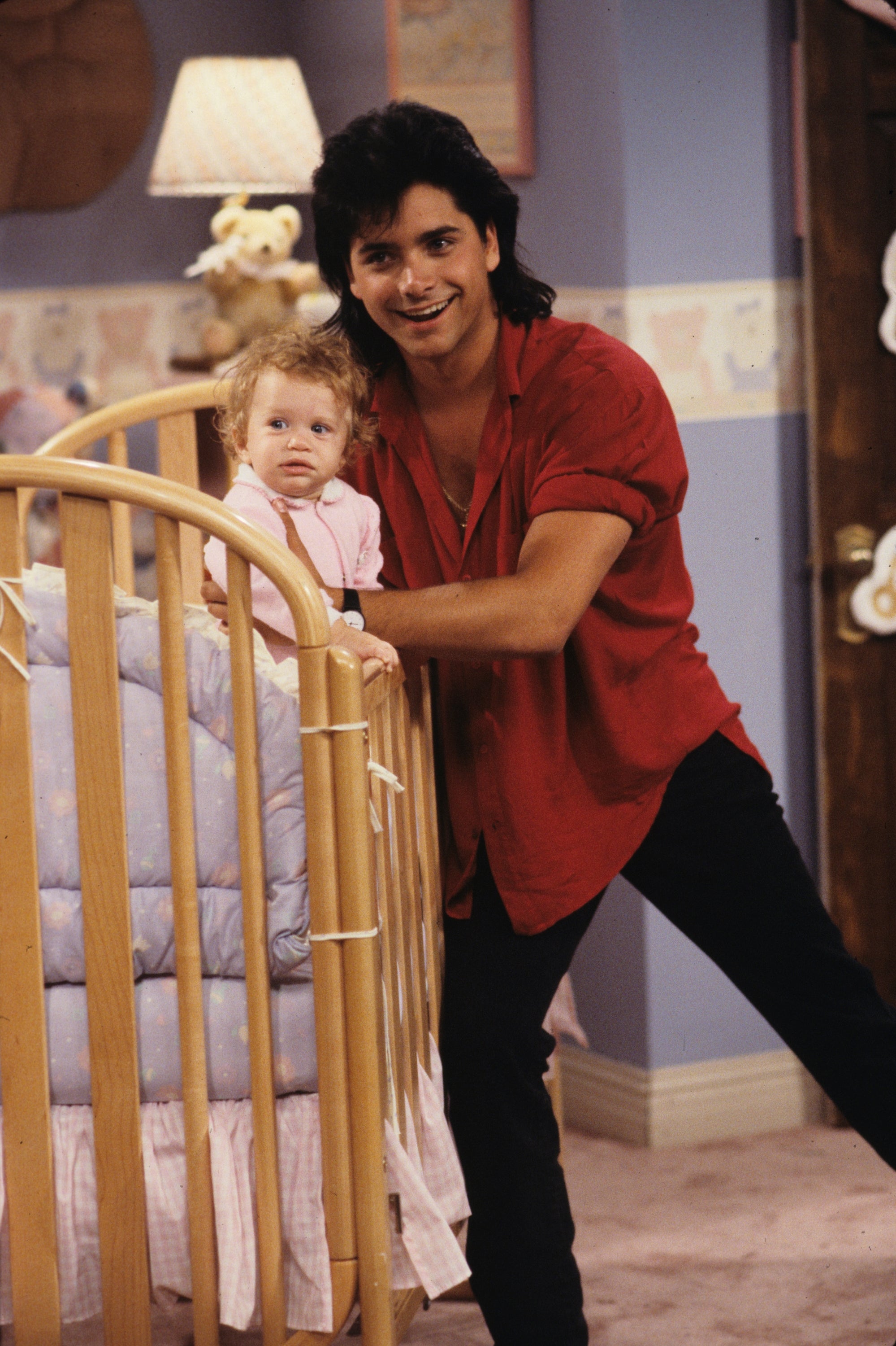 Person in a casual shirt leans on a crib, smiling, with a small child inside. The setting is a cozy room