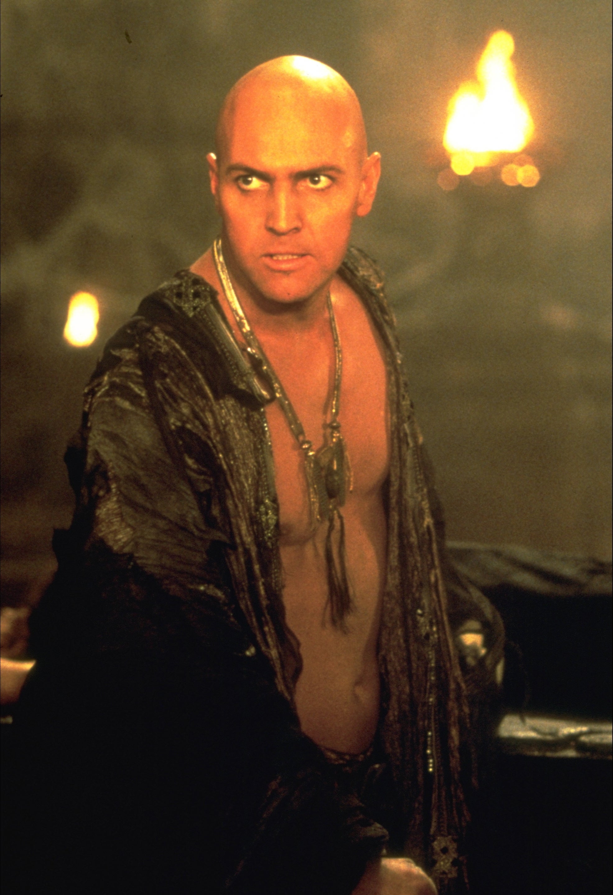 Man in ancient warrior attire with a bold expression, in a dimly lit setting with fire in the background