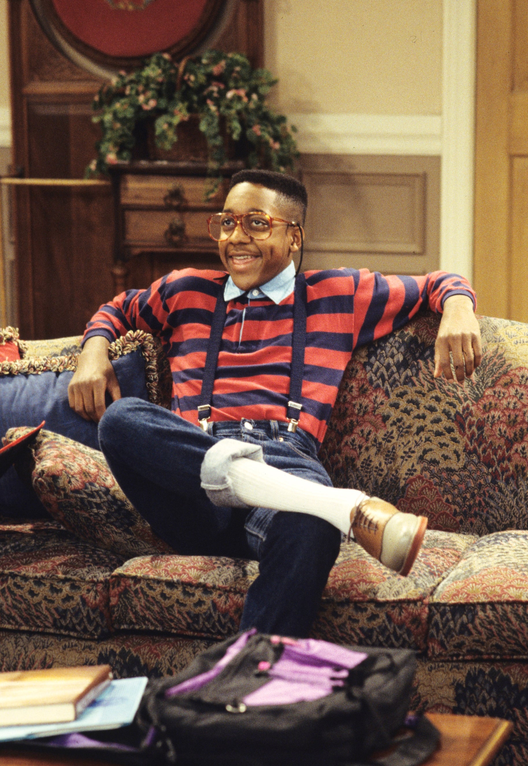 Person sitting on a patterned couch, wearing striped shirt, suspenders, and glasses, smiling broadly, evoking a 90s sitcom style