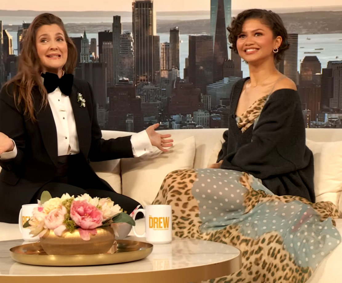 Two women are seated connected a speech amusement set. One wears a tuxedo; the different wears a patterned gown. They are engaged successful conversation, smiling