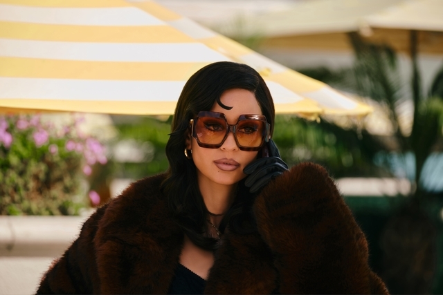 Person wearing oversized sunglasses and a luxurious faux fur overgarment nether a striped umbrella