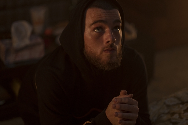 A idiosyncratic with a beard, wearing a hoodie, looks upward with clasped hands, appearing contemplative