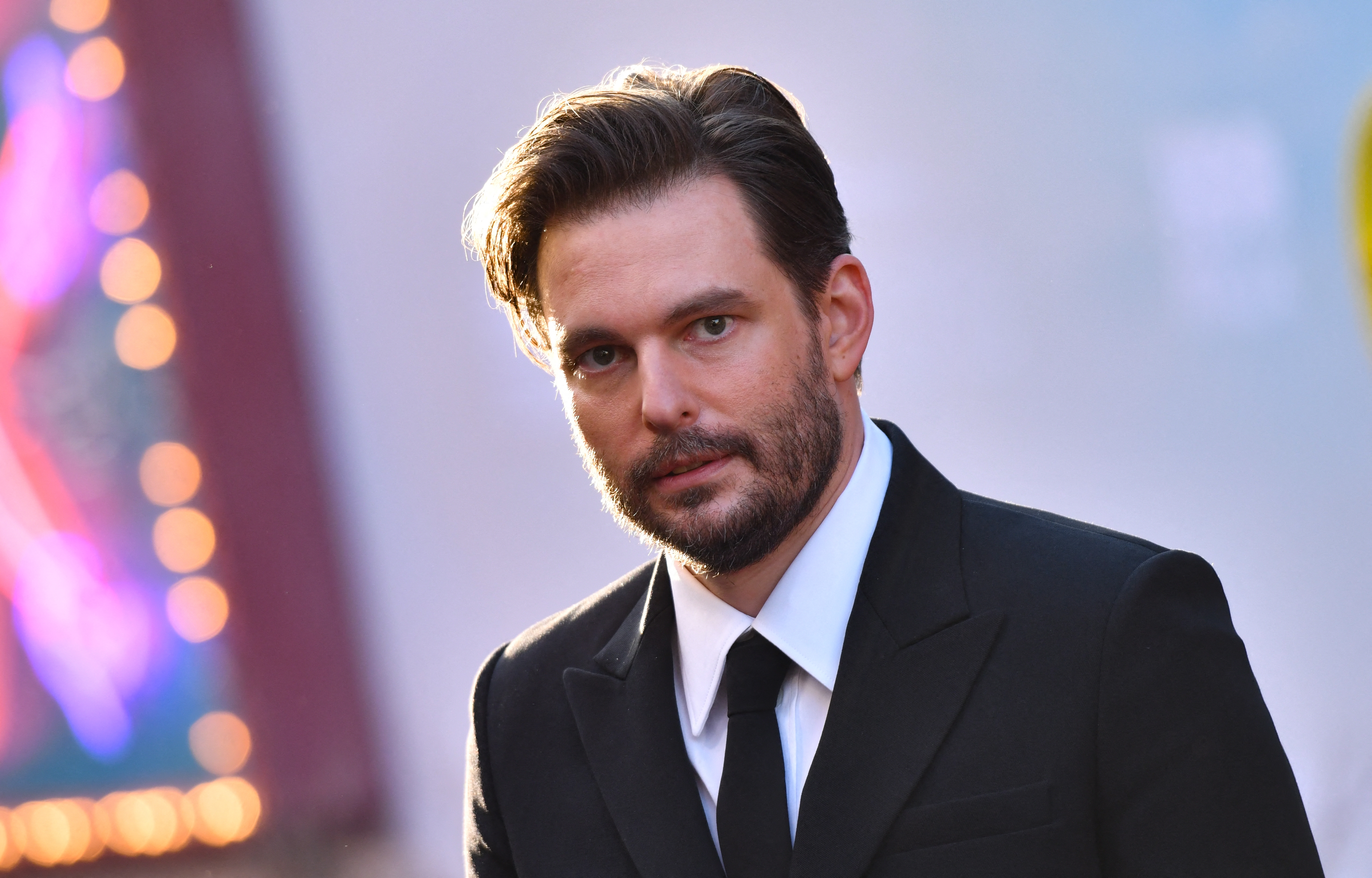 A idiosyncratic successful a ceremonial achromatic suit and necktie is posing connected the reddish carpet, looking towards the camera