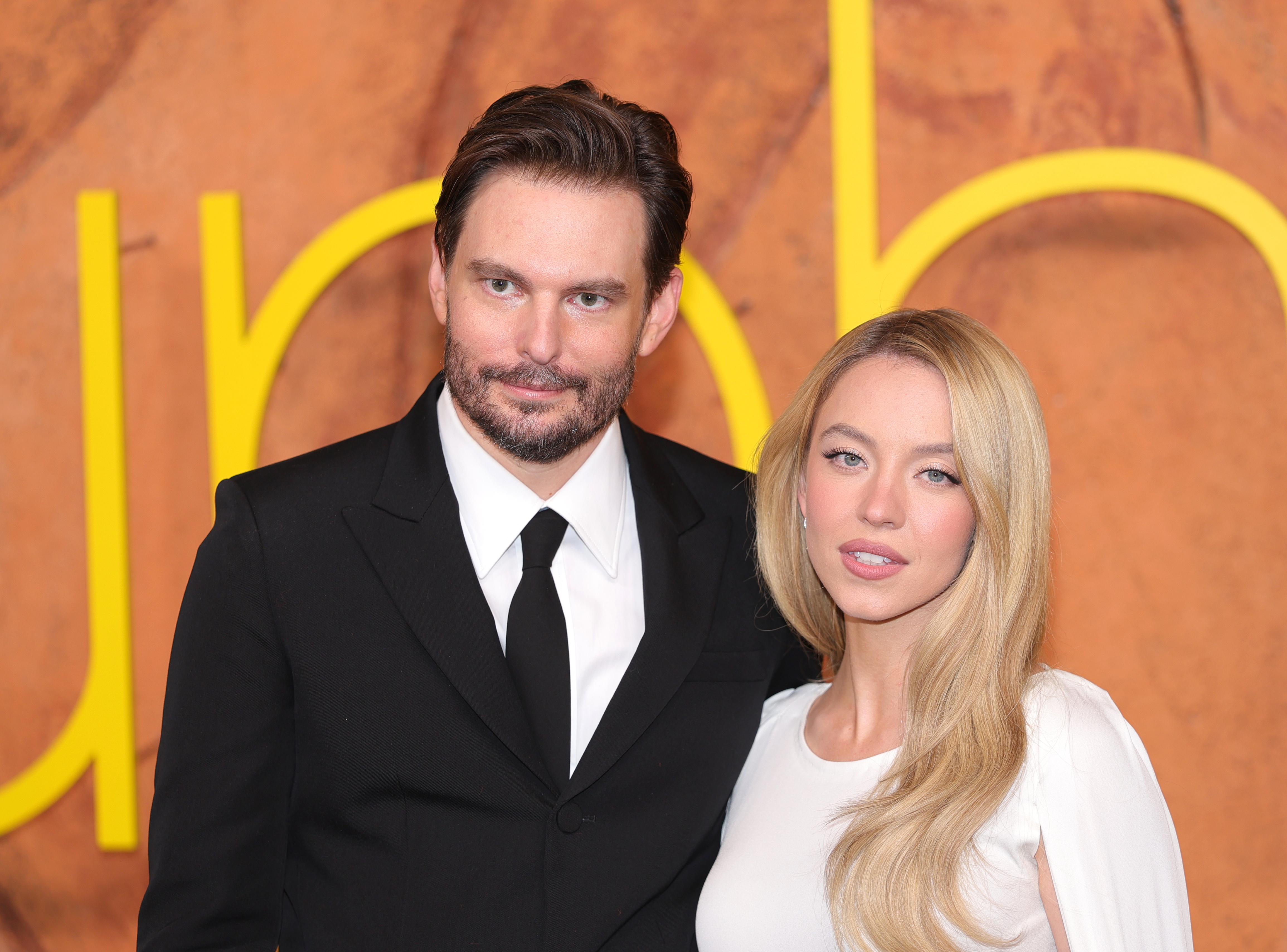 Two individuals connected a reddish carpet; 1 successful a achromatic suit and tie, the different successful a stylish achromatic dress, lasting successful beforehand of a ample substance display