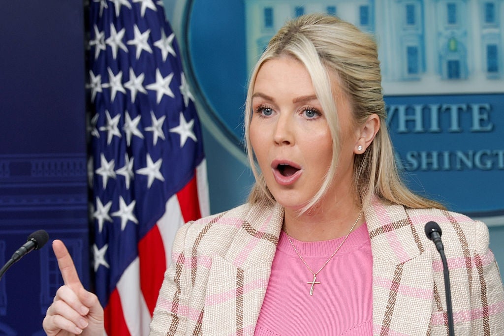Person som snakker lidenskapelig ved et pressepodium, iført en rutete blazer og korskjede, med et amerikansk flagg og Det hvite hus i bakgrunnen